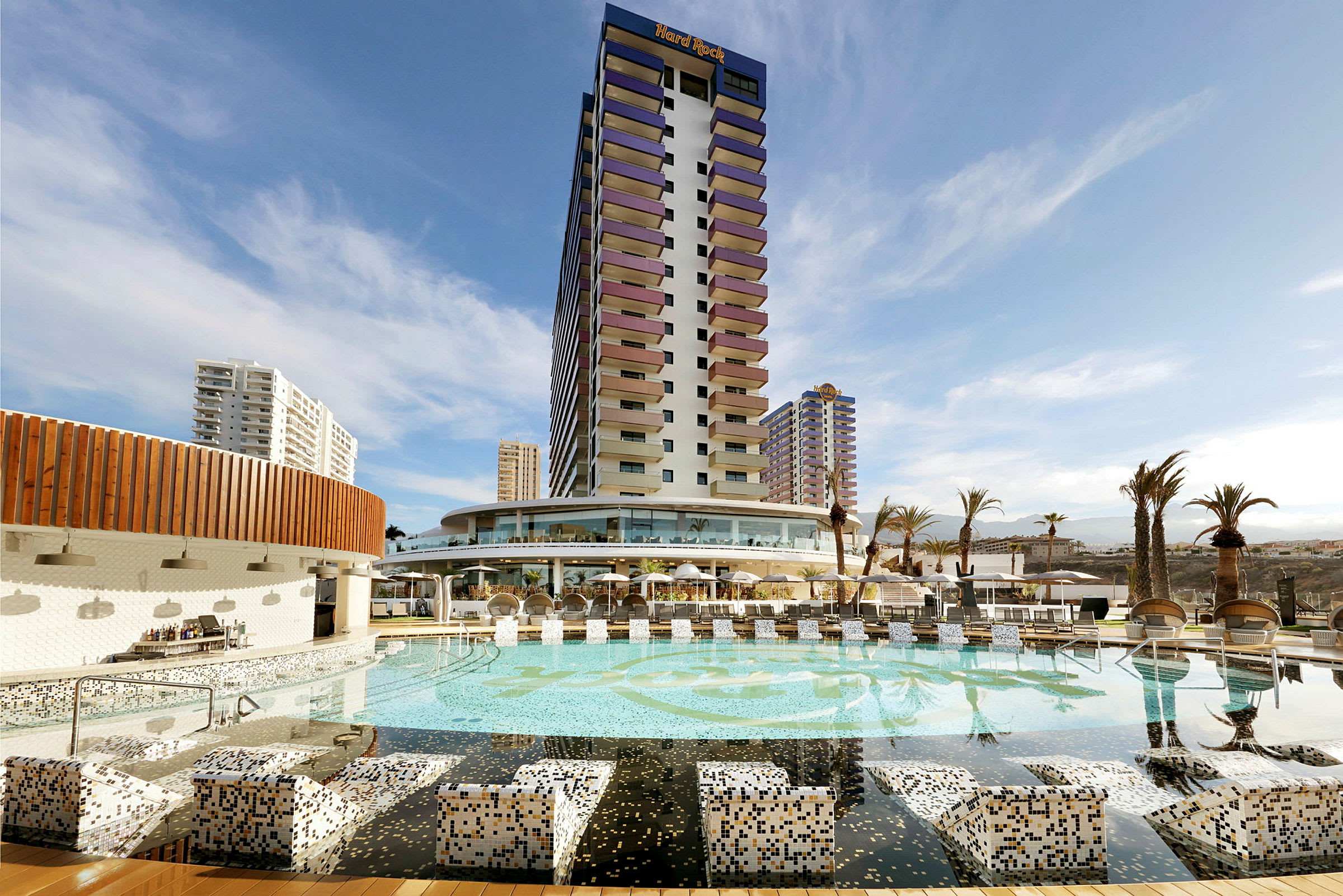 Ett hotell med en stor pool och omgivande byggnader under en blå himmel. (AI-genererad text)