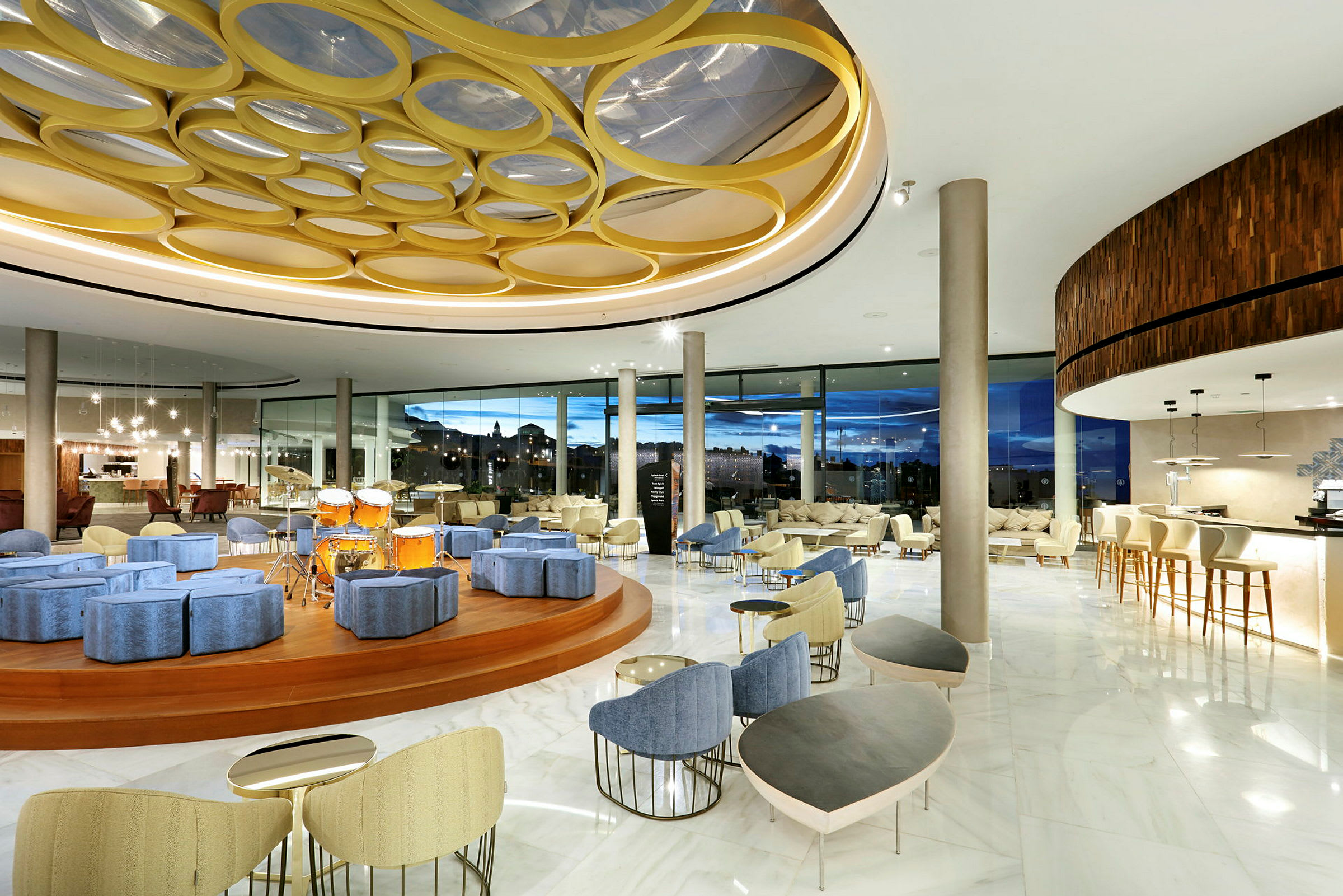Interiör av en modern hotellobby med sittgrupper, en scen med trummor och stora panoramafönster. (AI-genererad text)