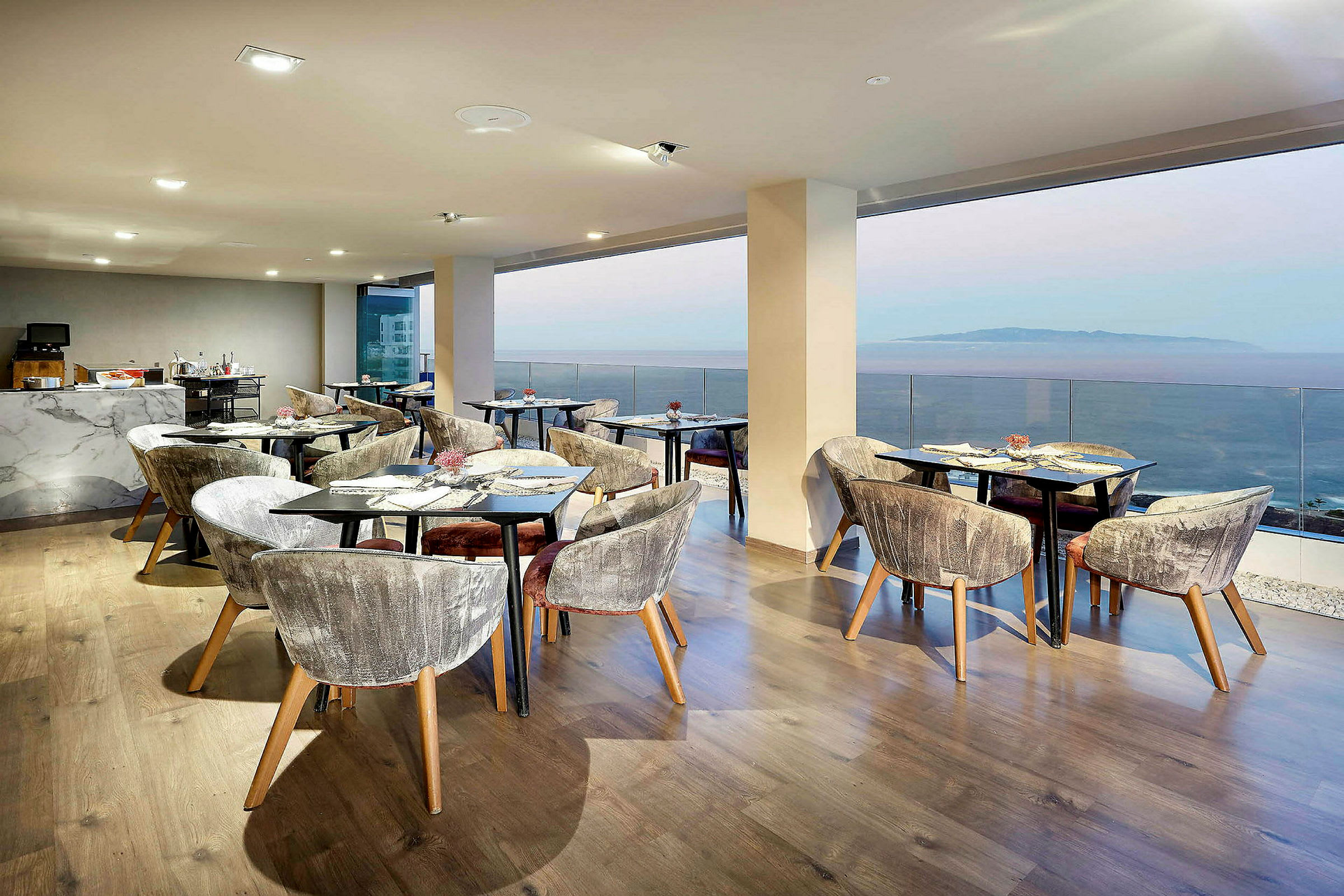 En restaurang med sittmöbler och bord framför stora fönster med utsikt över havet. (AI-genererad text)