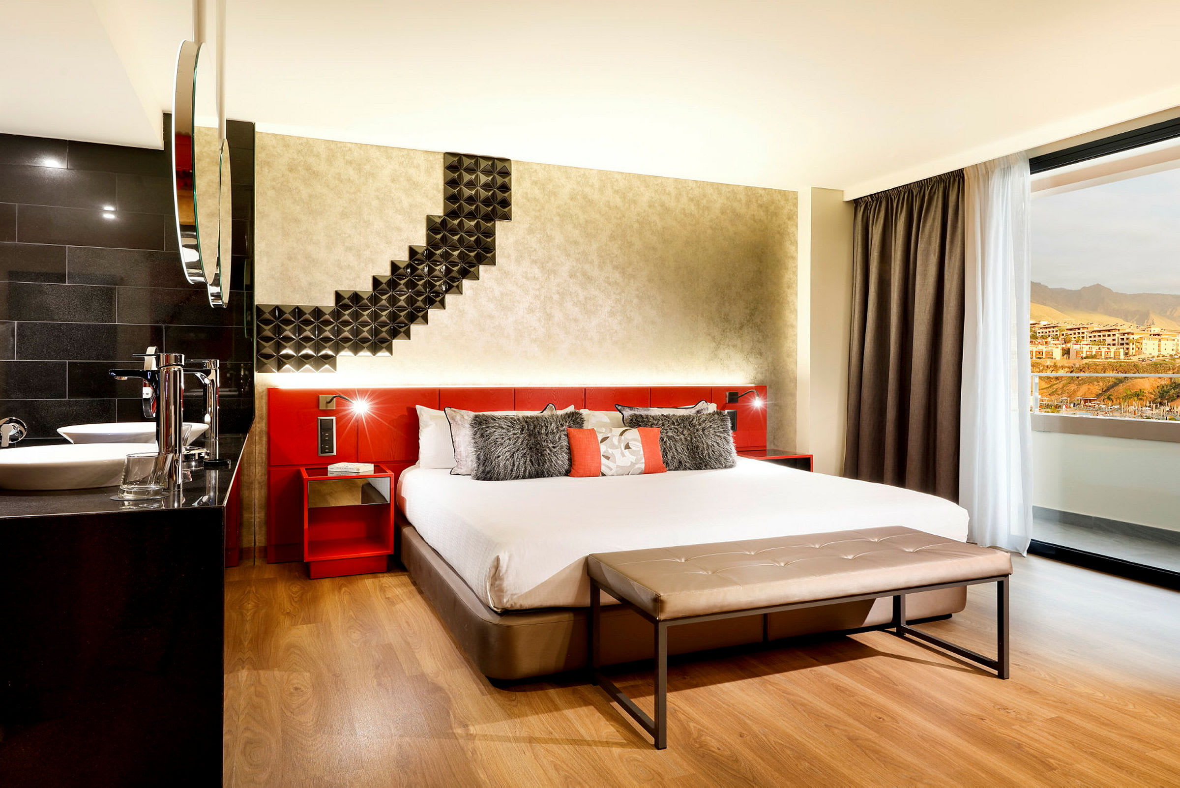 Ett hotellrum med en stor säng, modern inredning och en sittbänk framför sängen. Ett fönster visar utsikten mot berg och byggnader. (AI-genererad text)