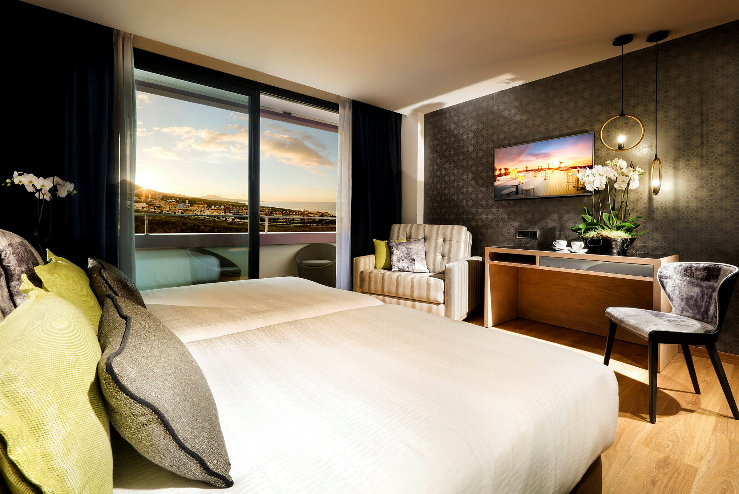 Hotellrum med dubbelsäng, fåtölj, skrivbord och utsikt mot solnedgången. (AI-genererad text)