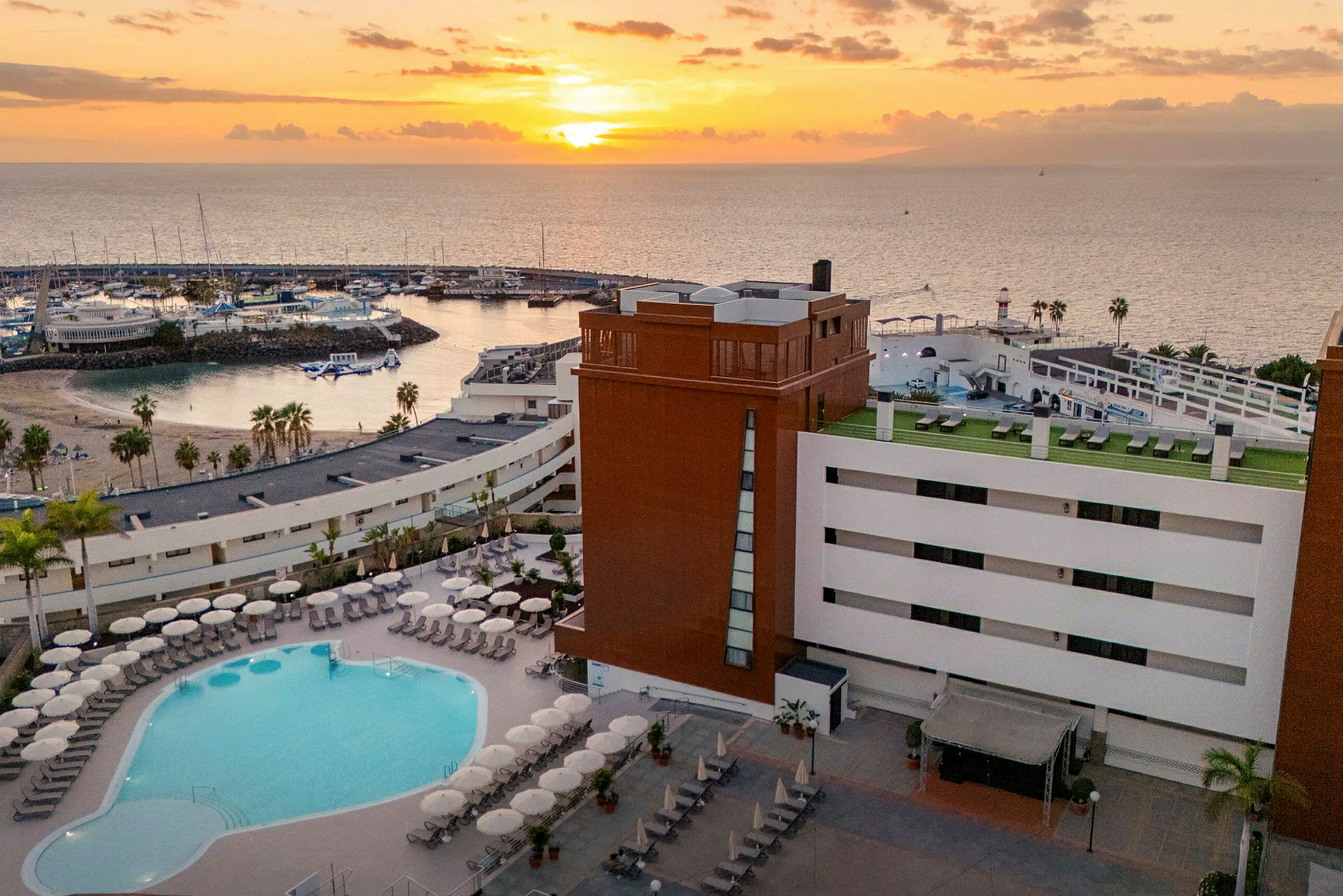 Bild av ett hotell med poolområde i förgrunden och en solnedgång över en marina i bakgrunden. (AI-genererad text)