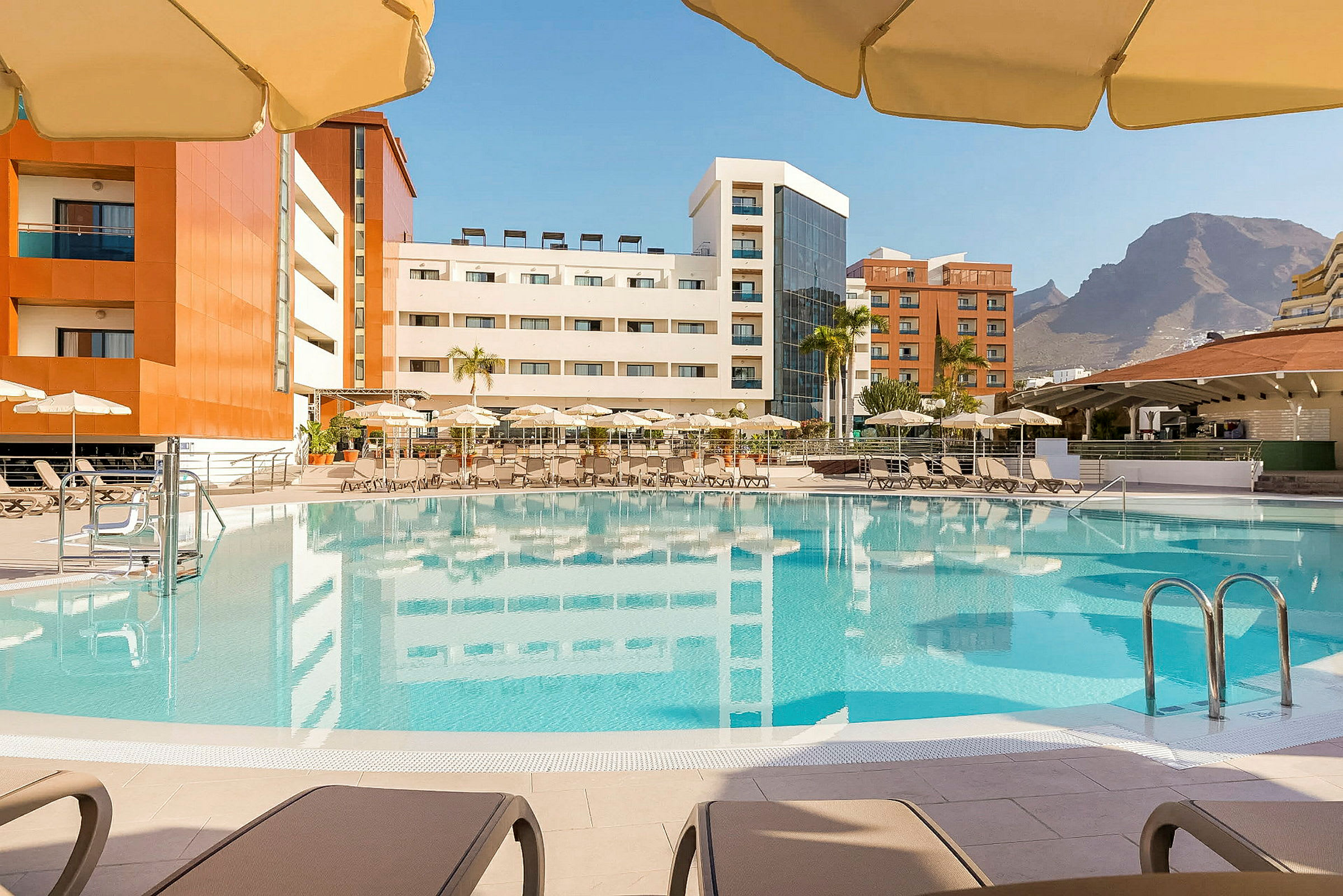 En pool med omgivande solstolar och parasoller, med hotellbyggnader i bakgrunden och berg i fjärran. (AI-genererad text)