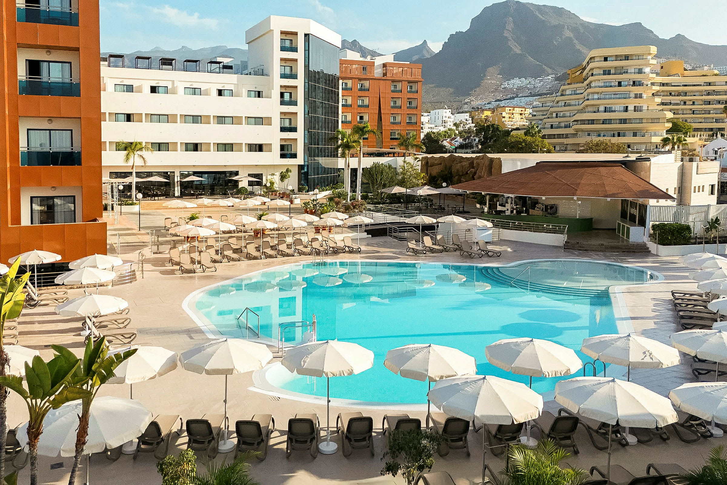 Hotell med poolområde, omgärdat av parasoller och solstolar, samt omgivande byggnader och berg i bakgrunden. (AI-genererad text)
