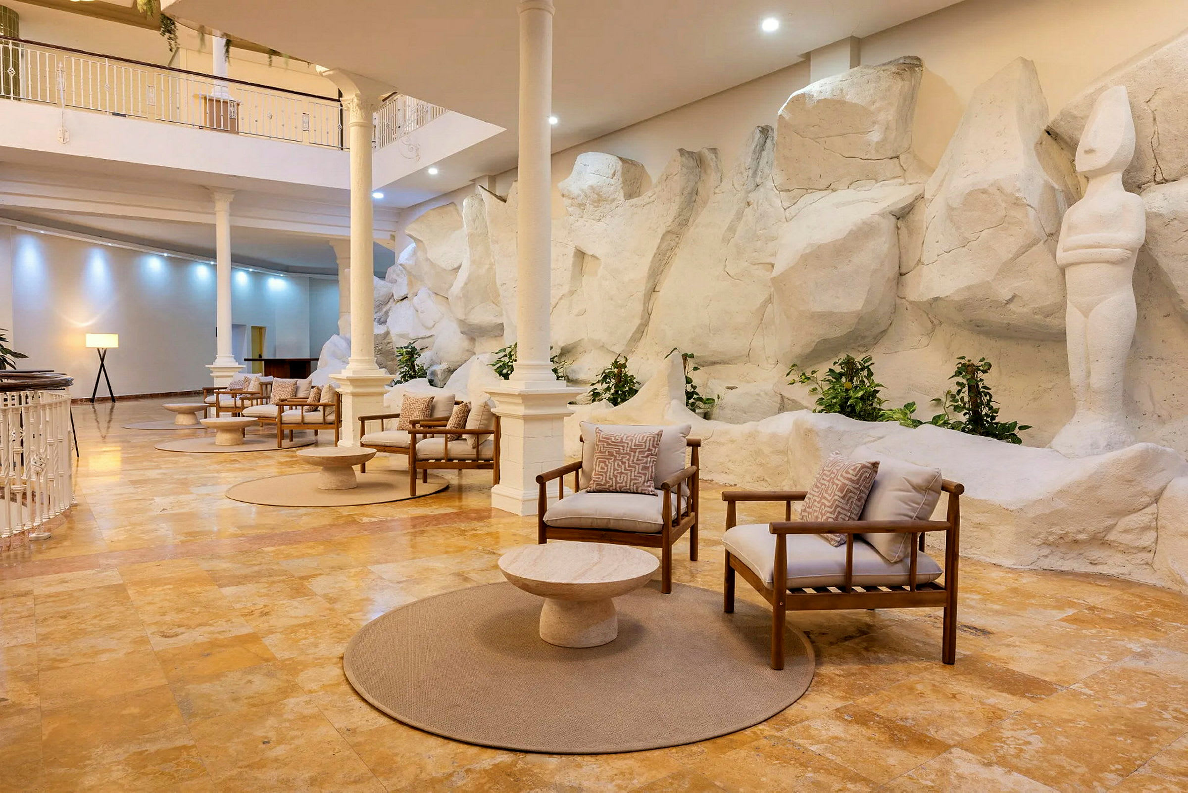 En elegant lounge med trästolar och kuddar, placerade på en stengolv, omgiven av vita skulpturer och stenväggar, under en öppen inomhusmiljö. (AI-genererad text)