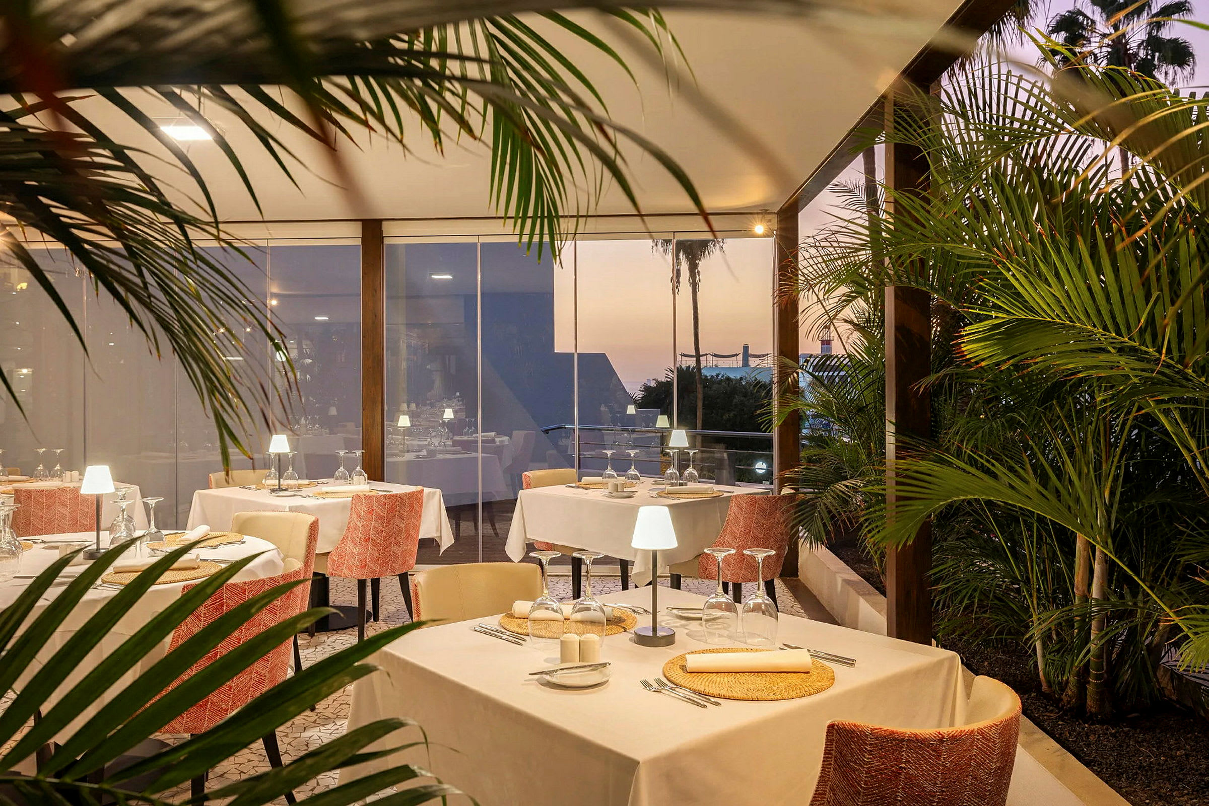En elegant restaurang med dukade bord inomhus och omgiven av grönska, med solnedgång i bakgrunden. (AI-genererad text)