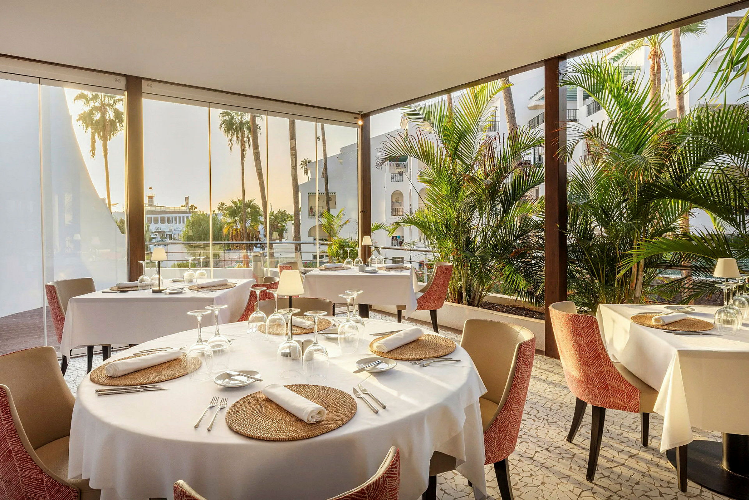 En restaurang med dukade bord, stora fönster och utsikt över palmer. (AI-genererad text)