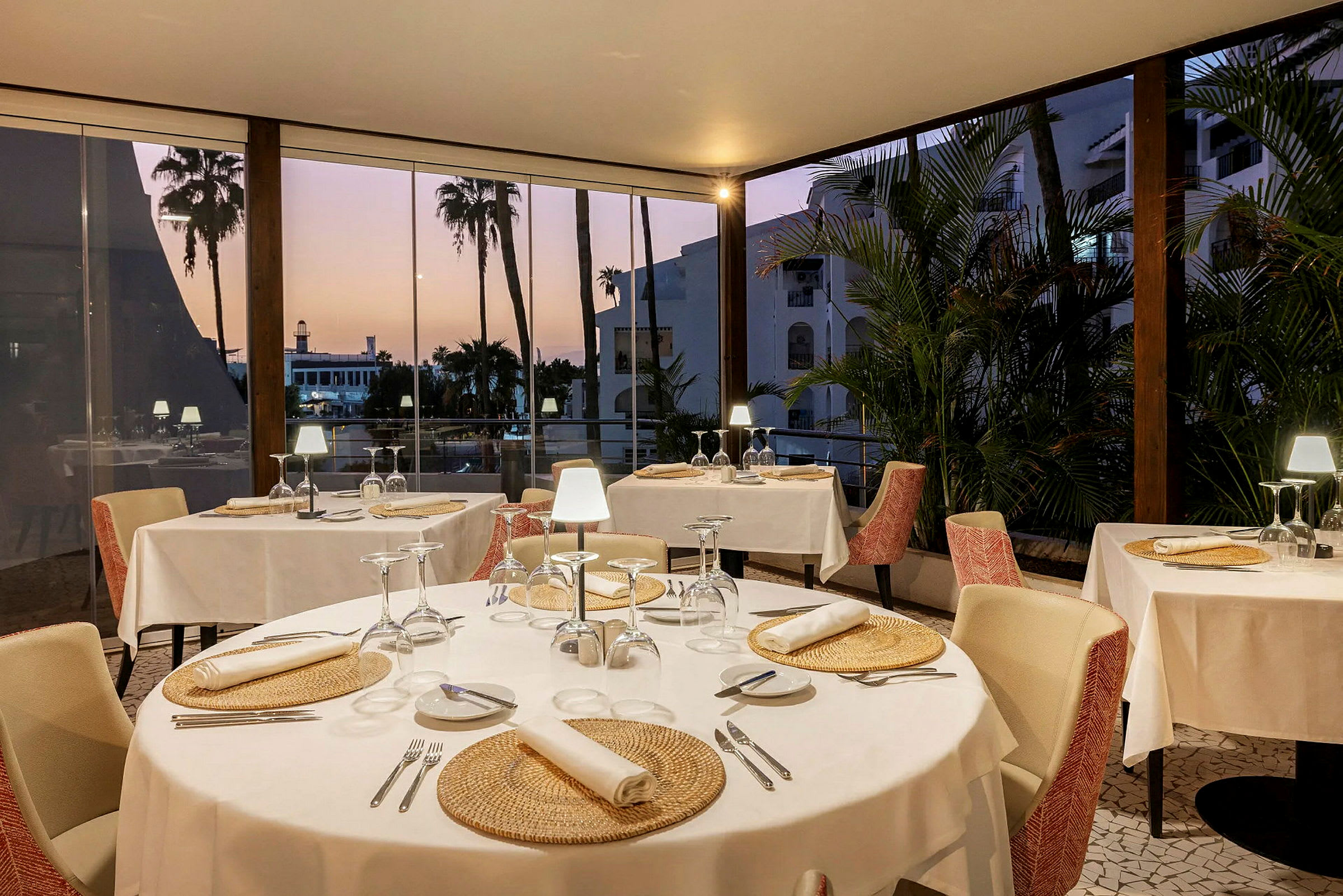 Inne i en restaurang med dukade bord och utsikt över palmer genom stora fönster. (AI-genererad text)