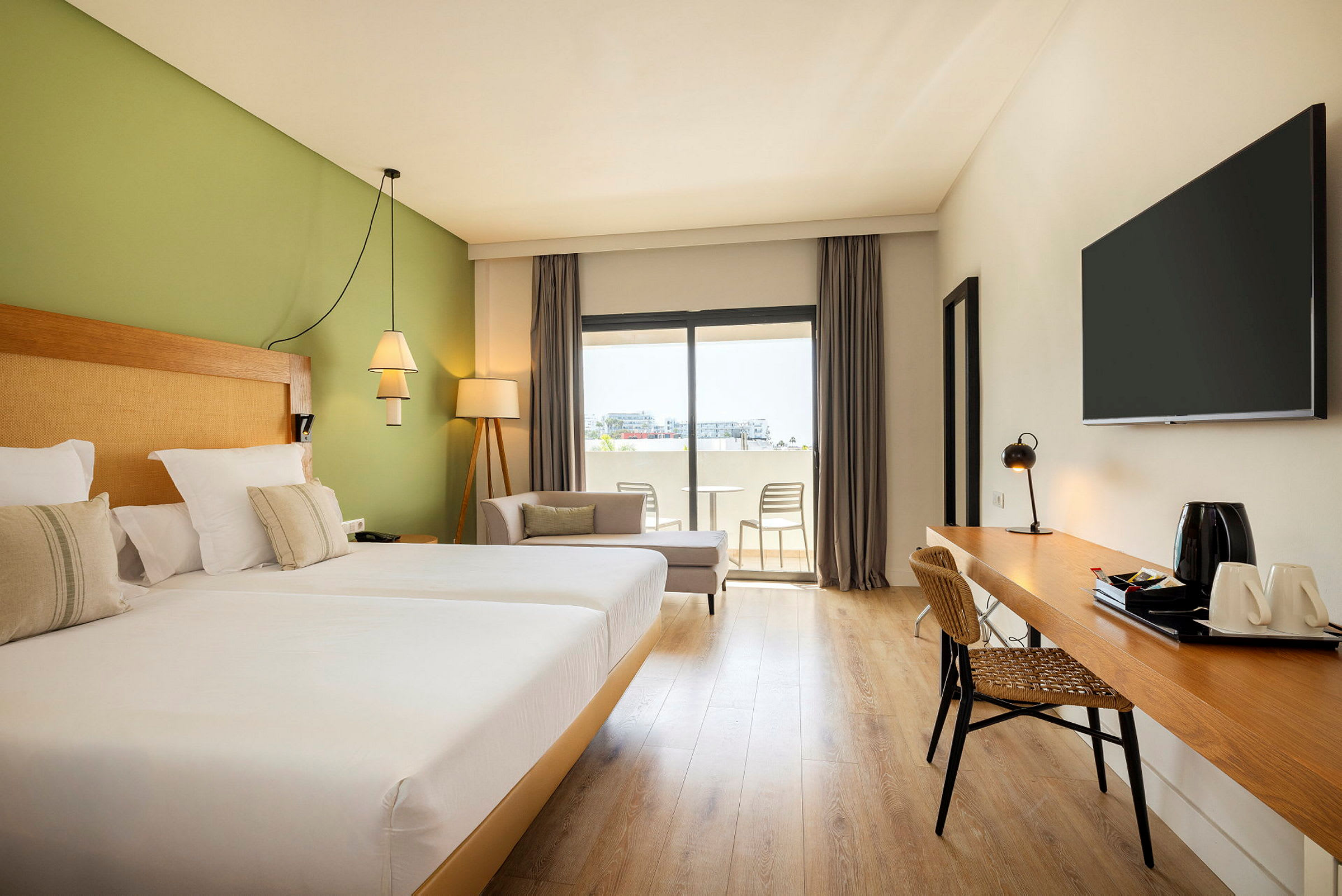 Ett hotellrum med två sängar, gröna väggar, skrivbord och en sittgrupp bredvid ett fönster med utsikt över balkongen. (AI-genererad text)