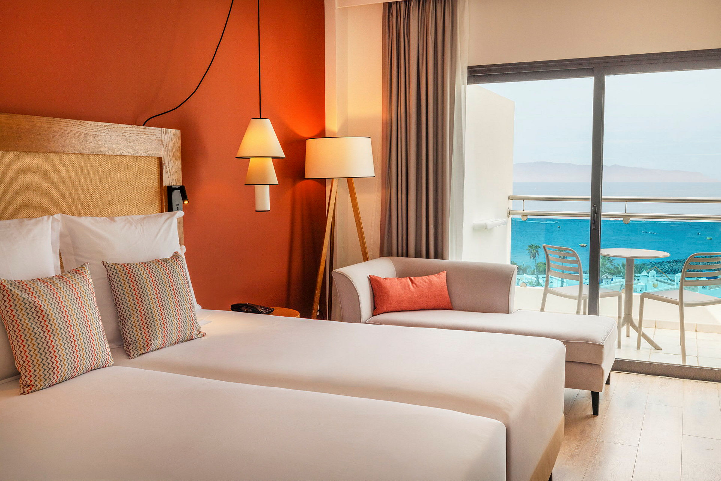 Ett hotellrum med dubbelsäng, dekorativa kuddar, en soffa och glasdörrar som leder till balkongen med utsikt över havet. (AI-genererad text)