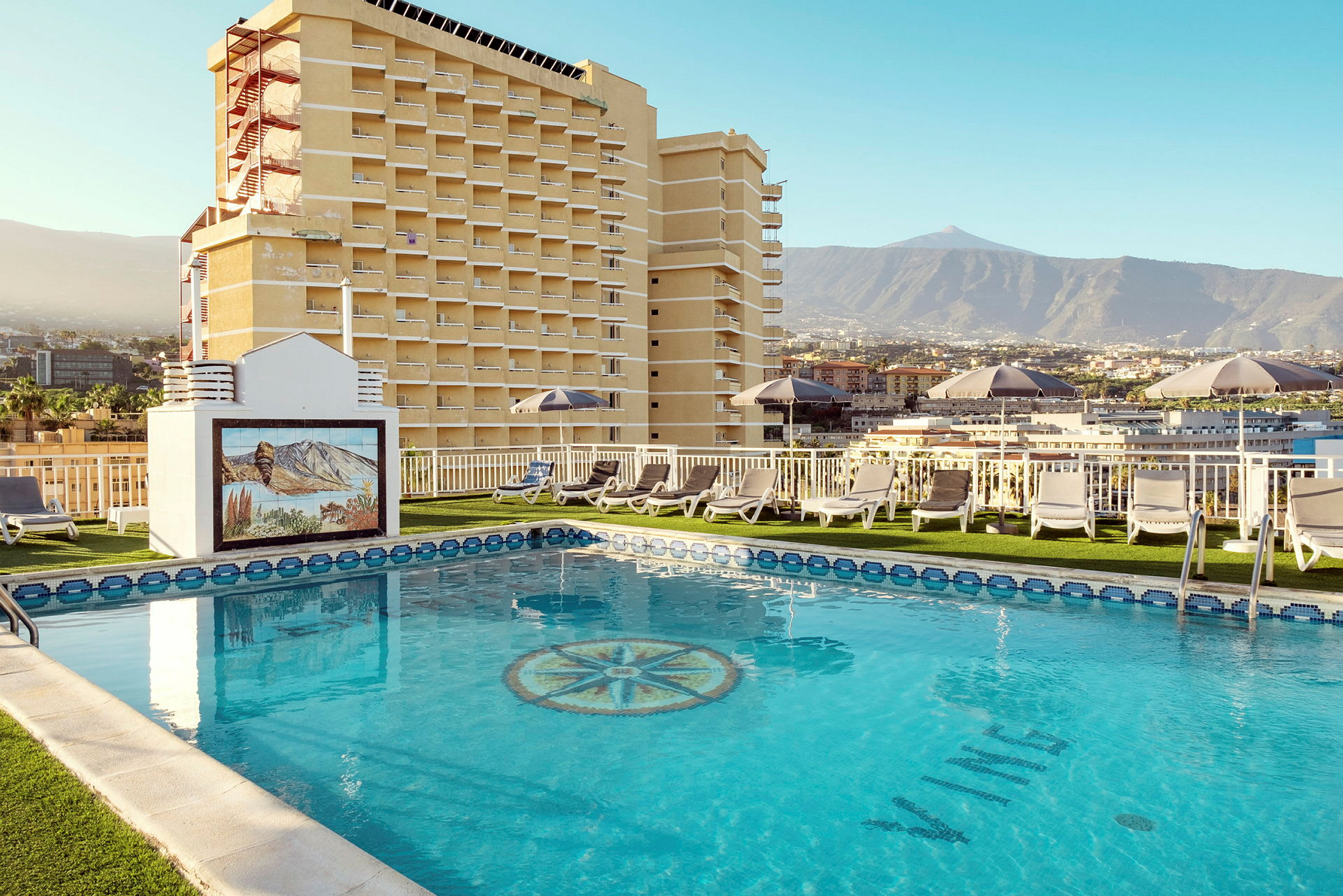 En hotellpool med solstolar och utsikt mot berg och byggnader. (AI-genererad text)