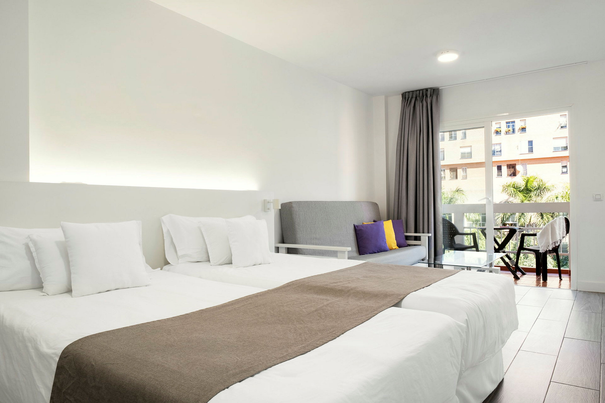 Ett hotellrum med två enkelsängar med vita sängkläder, en grå soffa med färgade kuddar och en balkong med utsikt. (AI-genererad text)