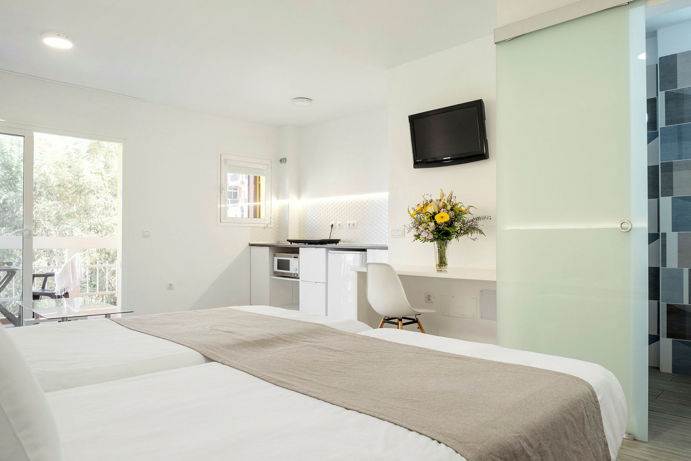 Ett hotellrum med en dubbelsäng, ljus inredning, ett kök, en balkong och ett bord med en vas blommor. (AI-genererad text)