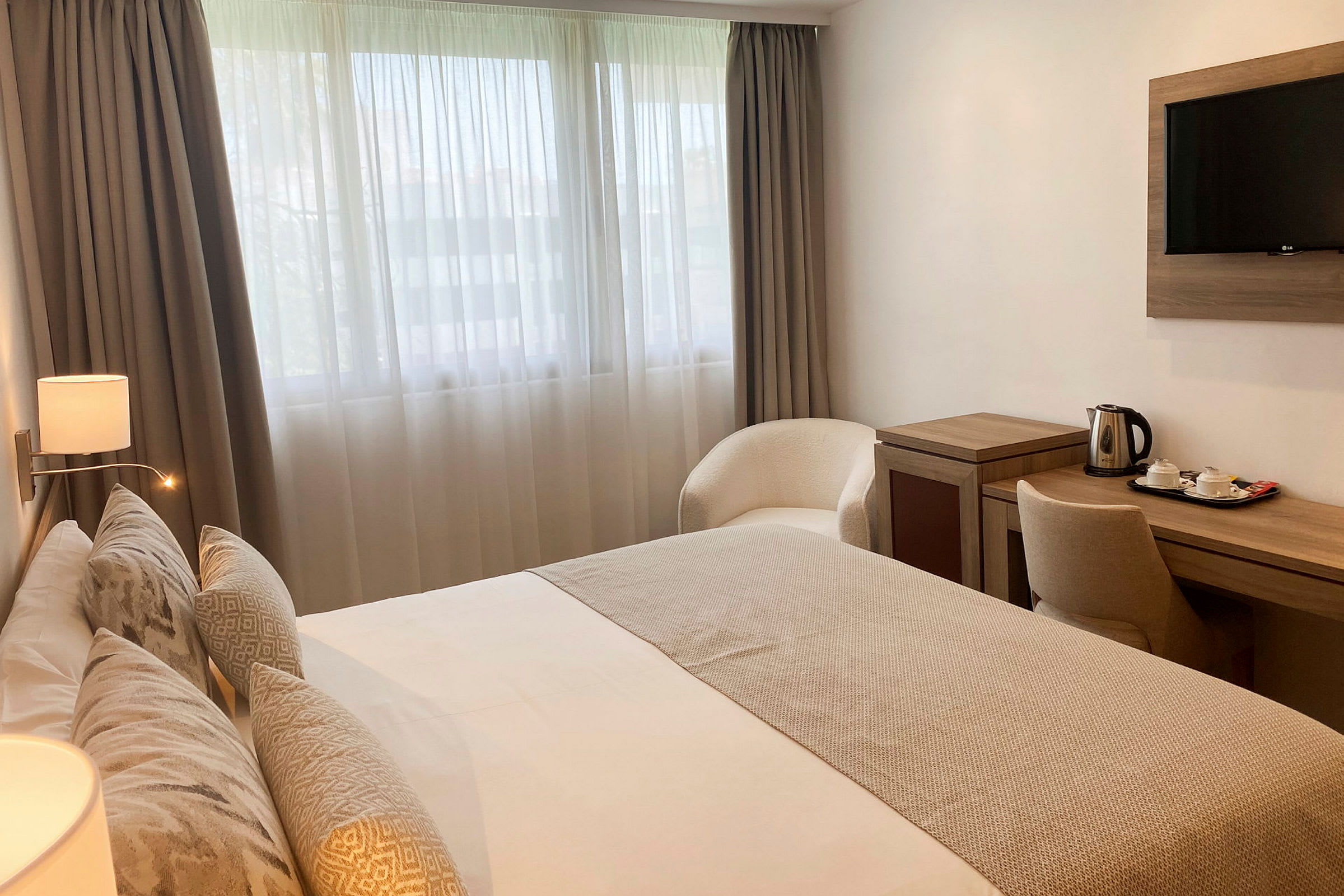 Hotellrum med en dubbelsäng, skrivbord med en vattenkokare och tefatsset, samt en fåtölj placerad nära fönstret. (AI-genererad text)