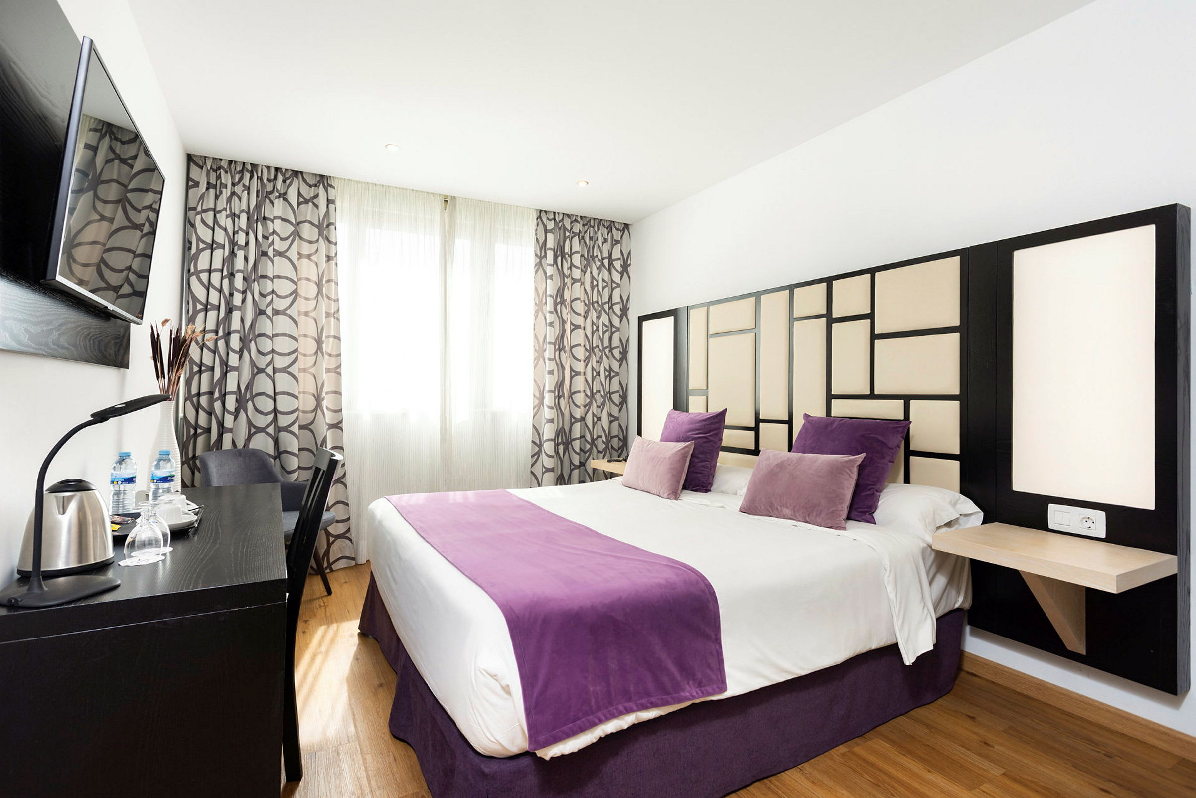 Ett hotellrum med en dubbelsäng, dekorerad med lila kuddar och överkast och en modern väggdesign. (AI-genererad text)
