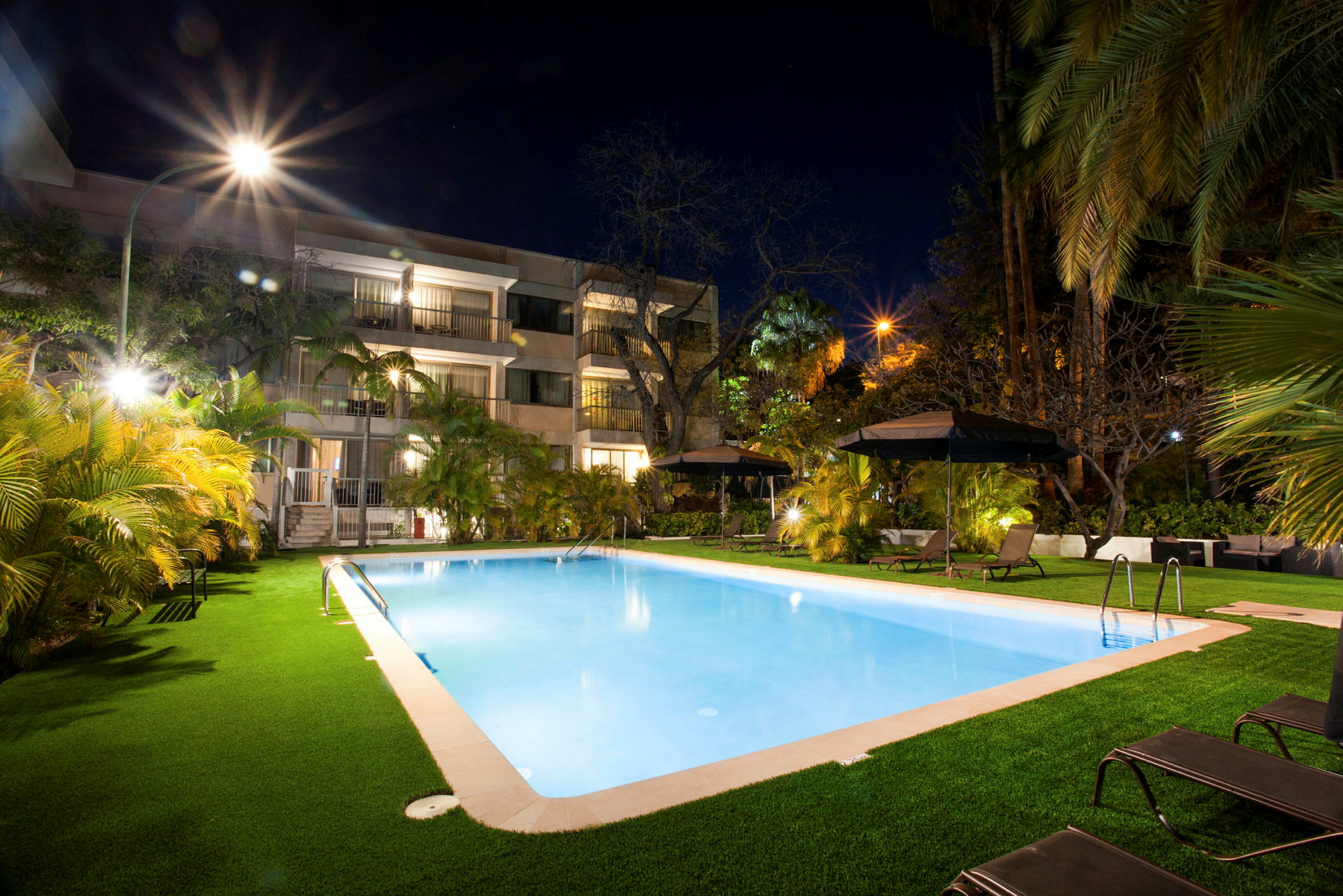En upplyst swimmingpool på kvällen vid ett hotell med omgivande vegetation och solstolar. (AI-genererad text)