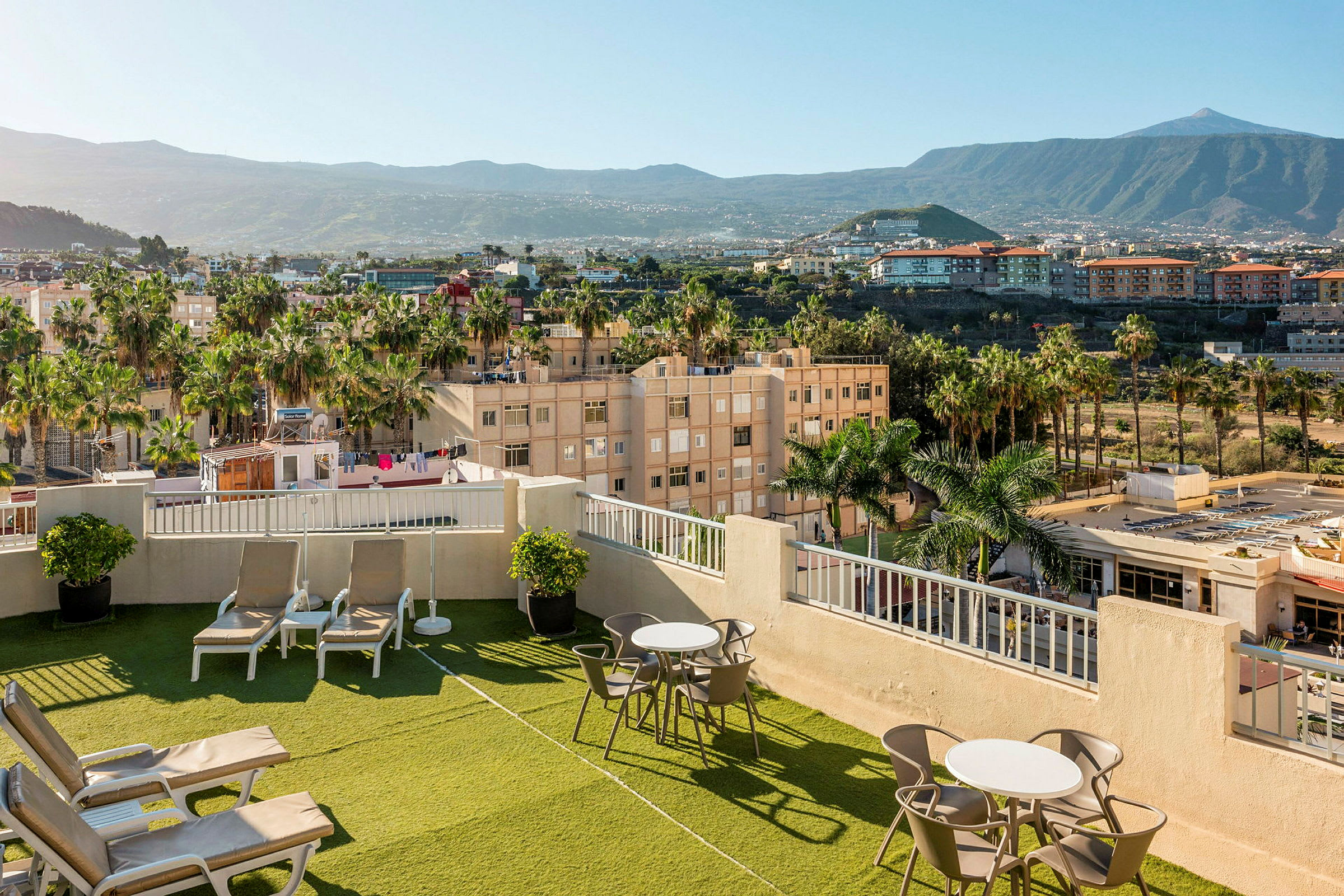 Bild av en hotellets takterrass med solstolar och bord, omgiven av gröna områden och palmer, med utsikt över omgivande stadsområde och berg. (AI-genererad text)