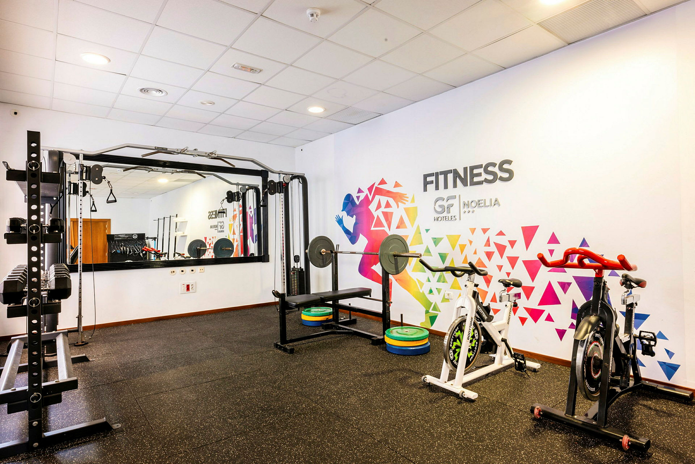 Bild på ett gym med träningsutrustning, inklusive skivstång och konditionscyklar, samt en vägg med färgglad dekoration. (AI-genererad text)