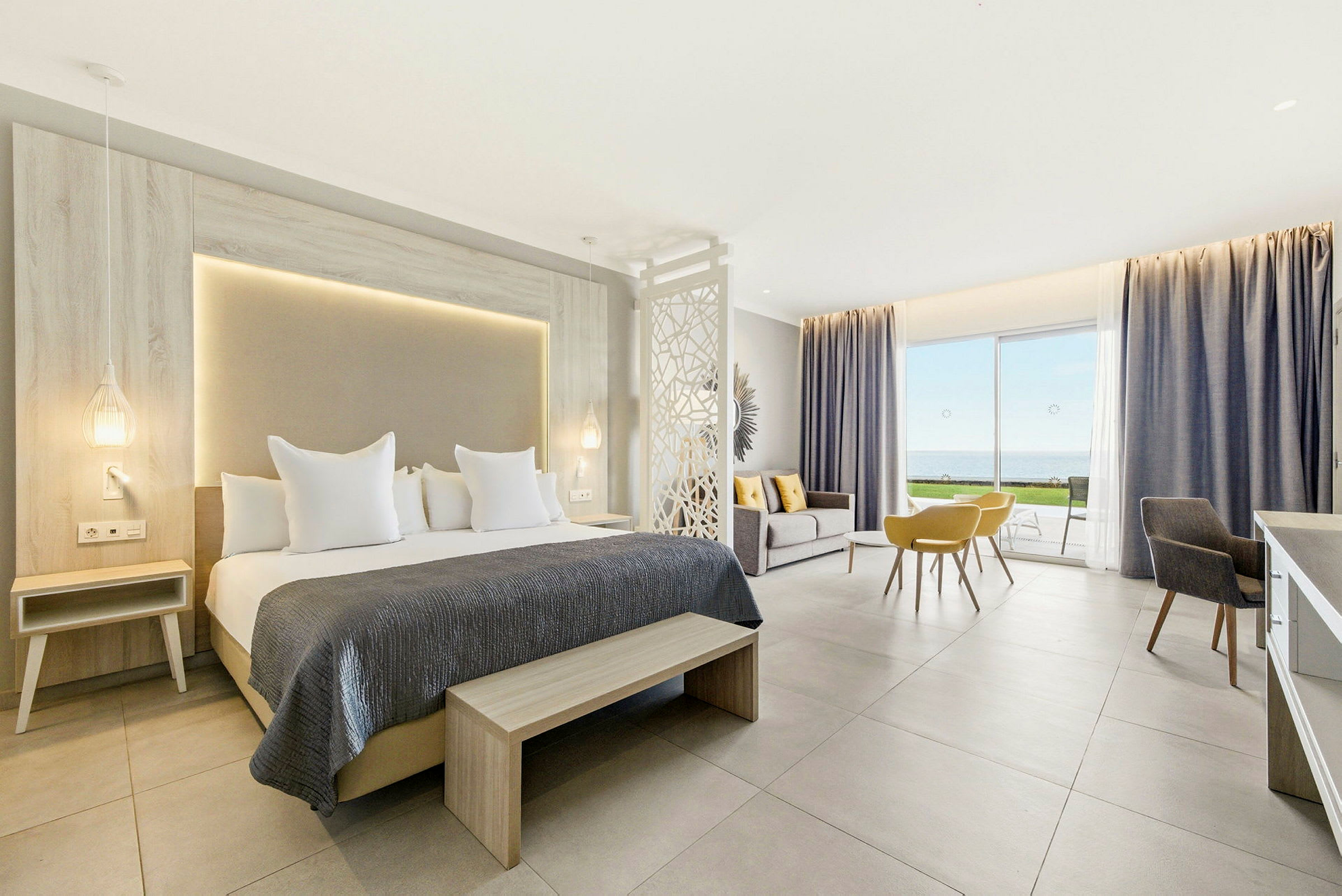 Ett modernt hotellrum med dubbelsäng, sittgrupp och utsikt över havet genom stora glasdörrar. (AI-genererad text)