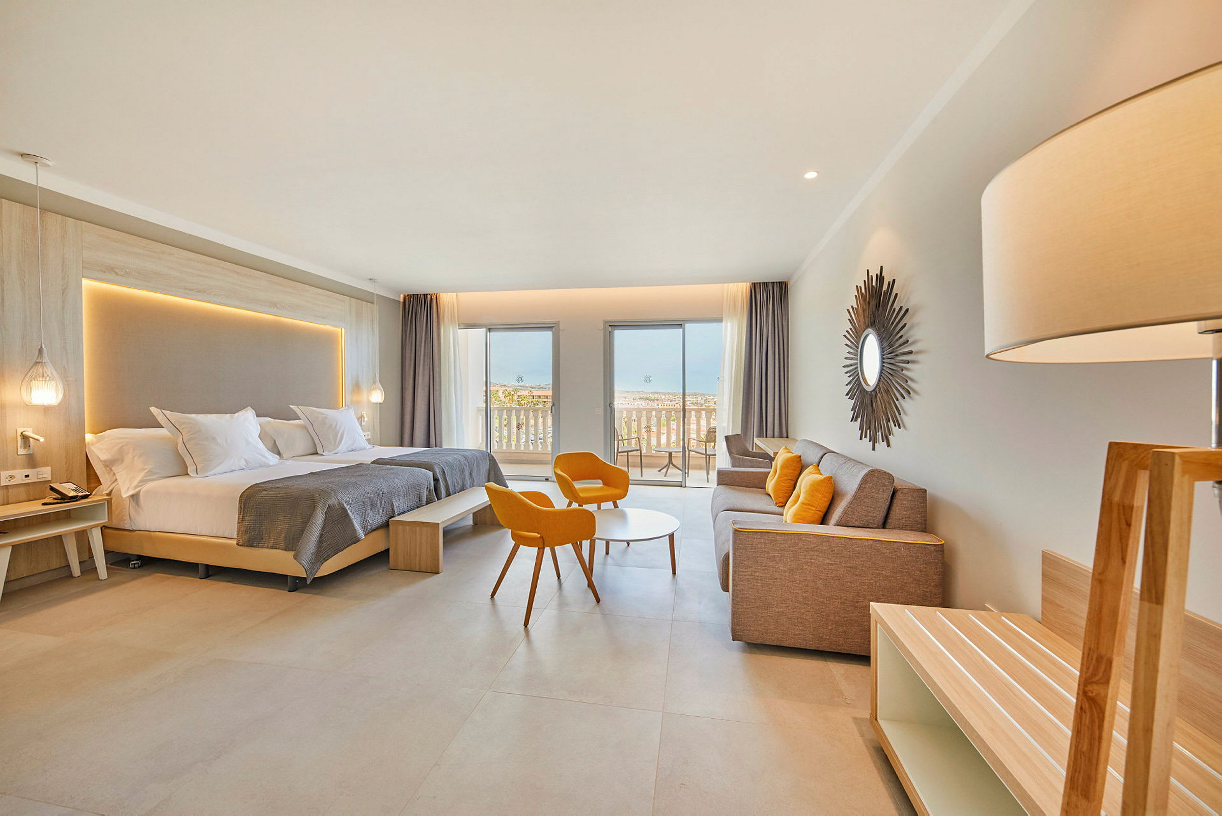 Ett modernt hotellrum med dubbelsäng, sittgrupp med orange stolar, en soffa, och en balkong som vetter mot havet. (AI-genererad text)