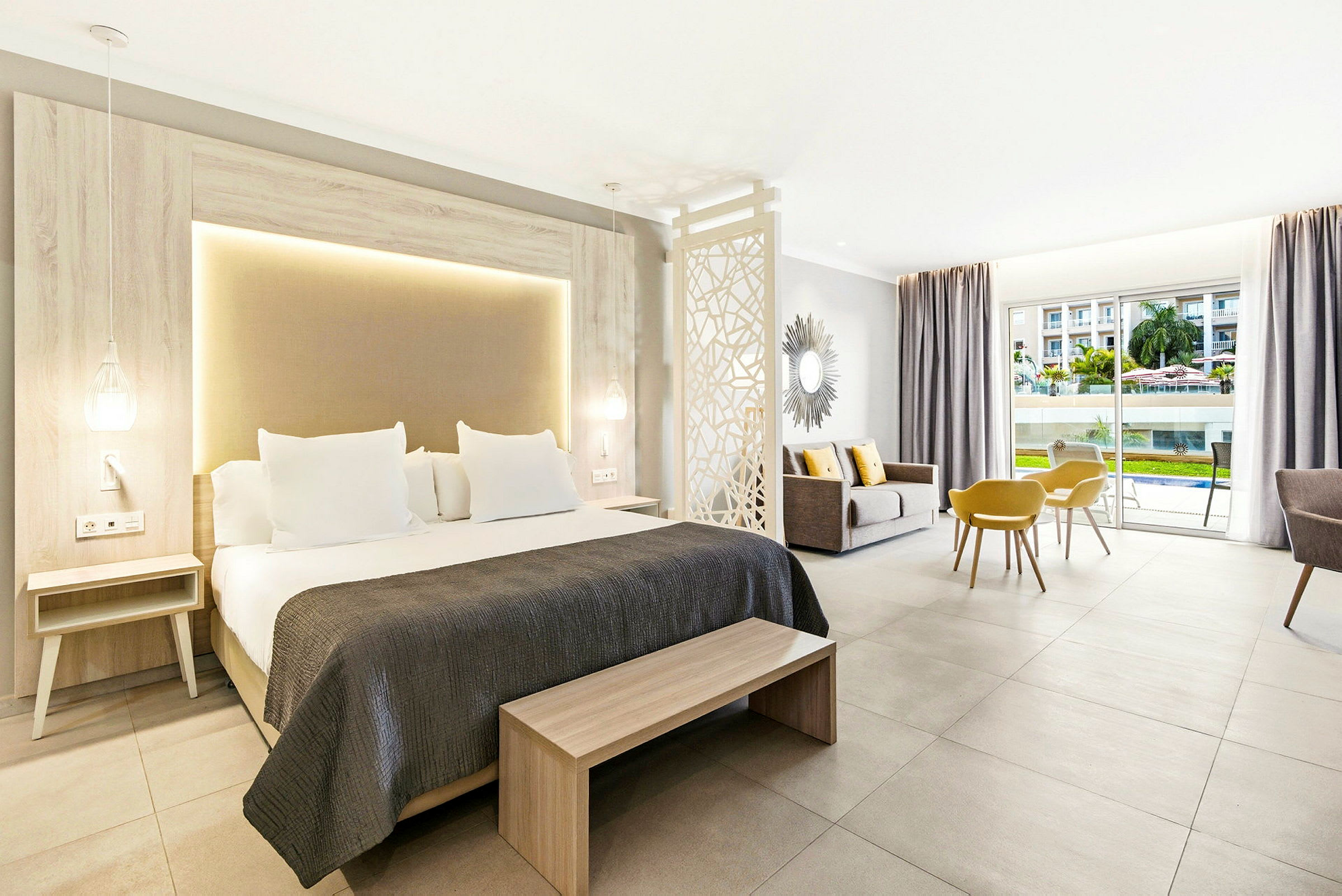 Ett modernt hotellrum med en dubbelsäng, sittgrupp och utsikt över poolområdet genom stora glasdörrar. (AI-genererad text)