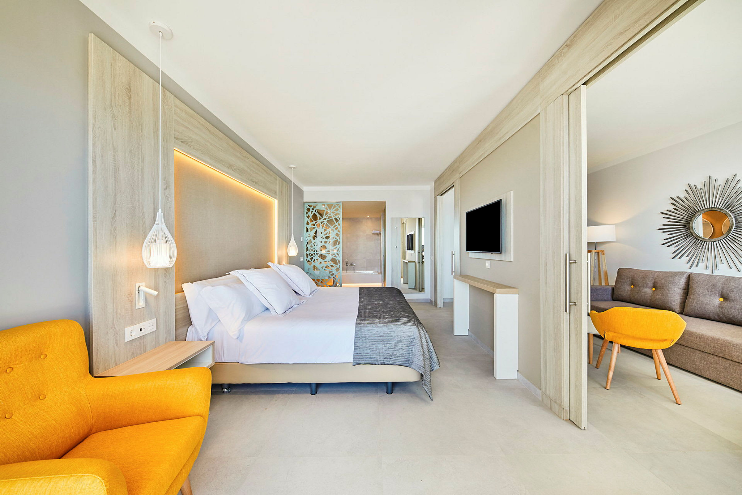 Ett modernt hotellrum med en stor säng, en gul fåtölj, samt en sittgrupp och platt-TV. Inredningen är ljus och stilren. (AI-genererad text)