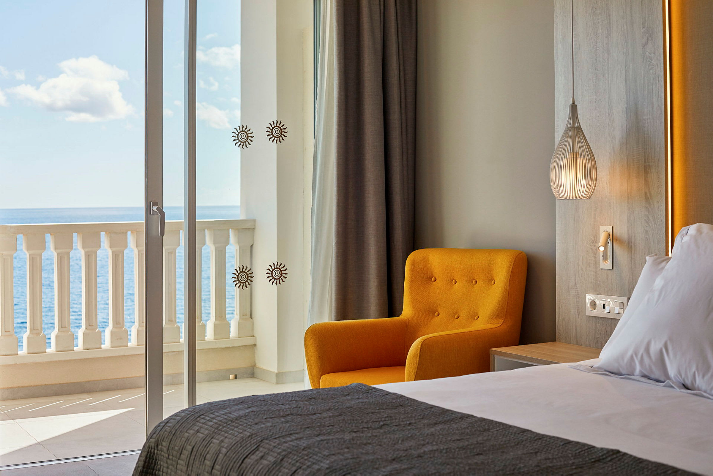 Ett hotellrum med havsutsikt. I förgrunden syns en bäddad säng, tillsammans med en gul stol och en modern lampa. (AI-genererad text)