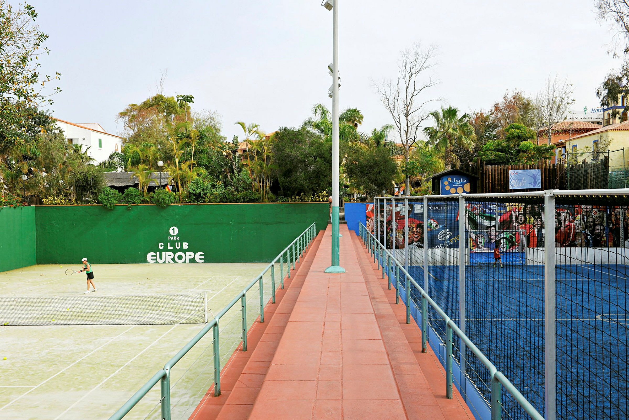 En grön tennisbana till vänster och en blå padelbana till höger, omgivna av träd och byggnader. (AI-genererad text)