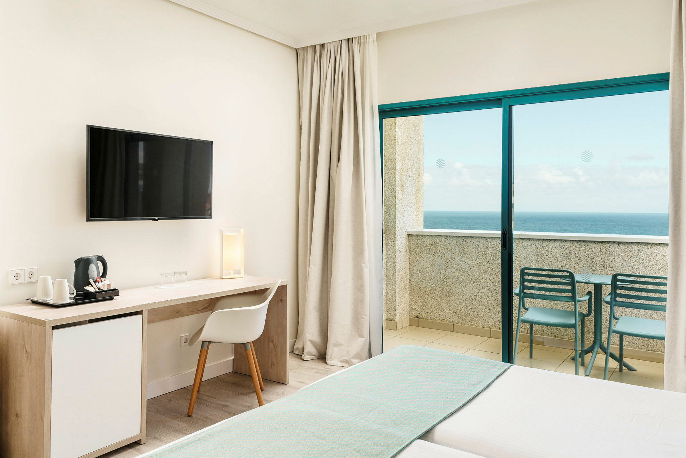 Interiör av ett hotellrum med en säng, skrivbord, TV och en balkong med utsikt över havet. (AI-genererad text)