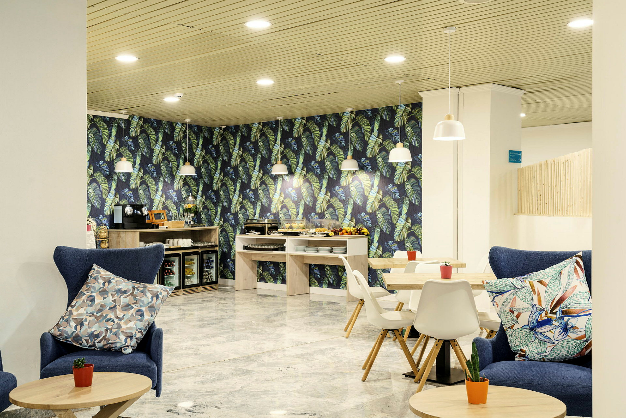 Ett caféutrymme med blåa fåtöljer, träbord, vita stolar och en dekorativ vägg med bladmotiv. Ett matbord och kaffe- samt dryckesutrustning är synliga. (AI-genererad text)