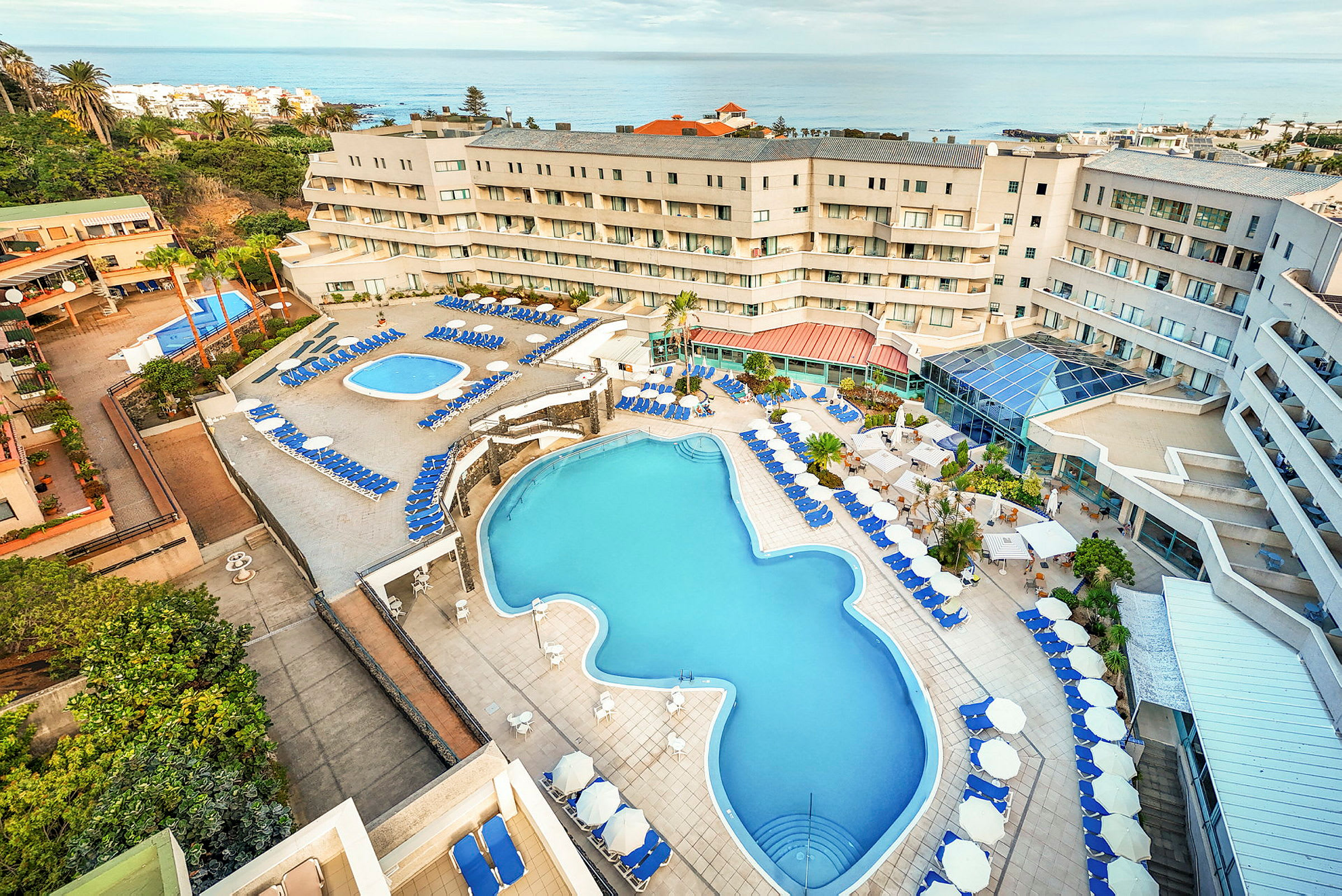 Ett hotell med poolområde som inkluderar två pooler, solstolar och parasoller, omgiven av grönska och med havet i bakgrunden. (AI-genererad text)