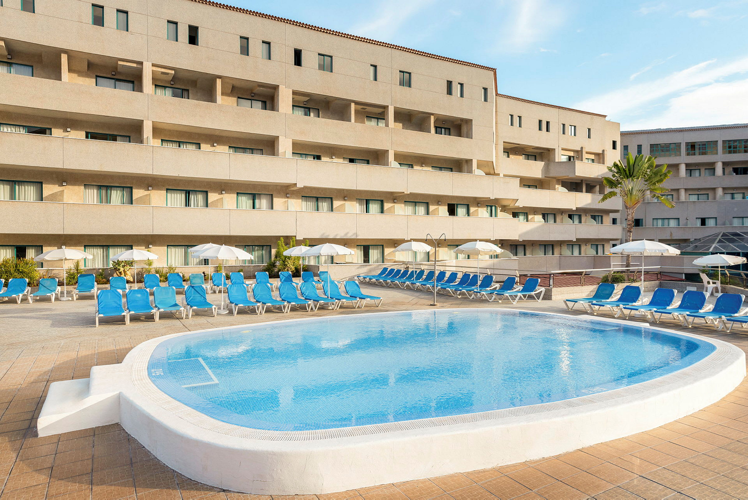 En pool med solstolar och parasoll framför en hotellbyggnad. (AI-genererad text)