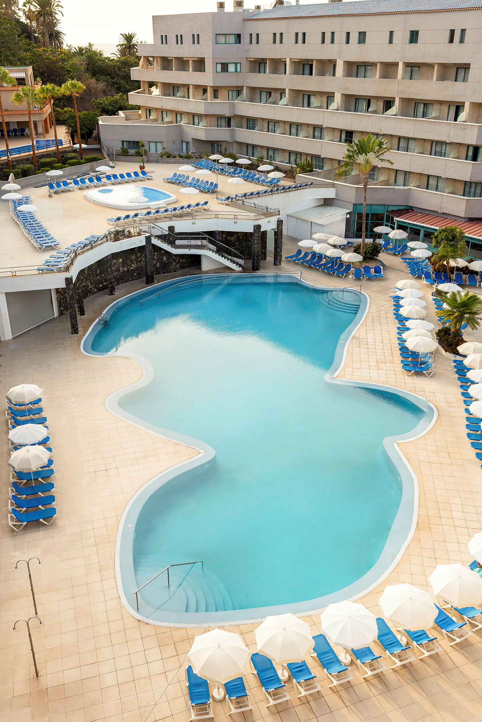En pool med blå solstolar och vita parasoll ligger framför en hotellbyggnad. (AI-genererad text)
