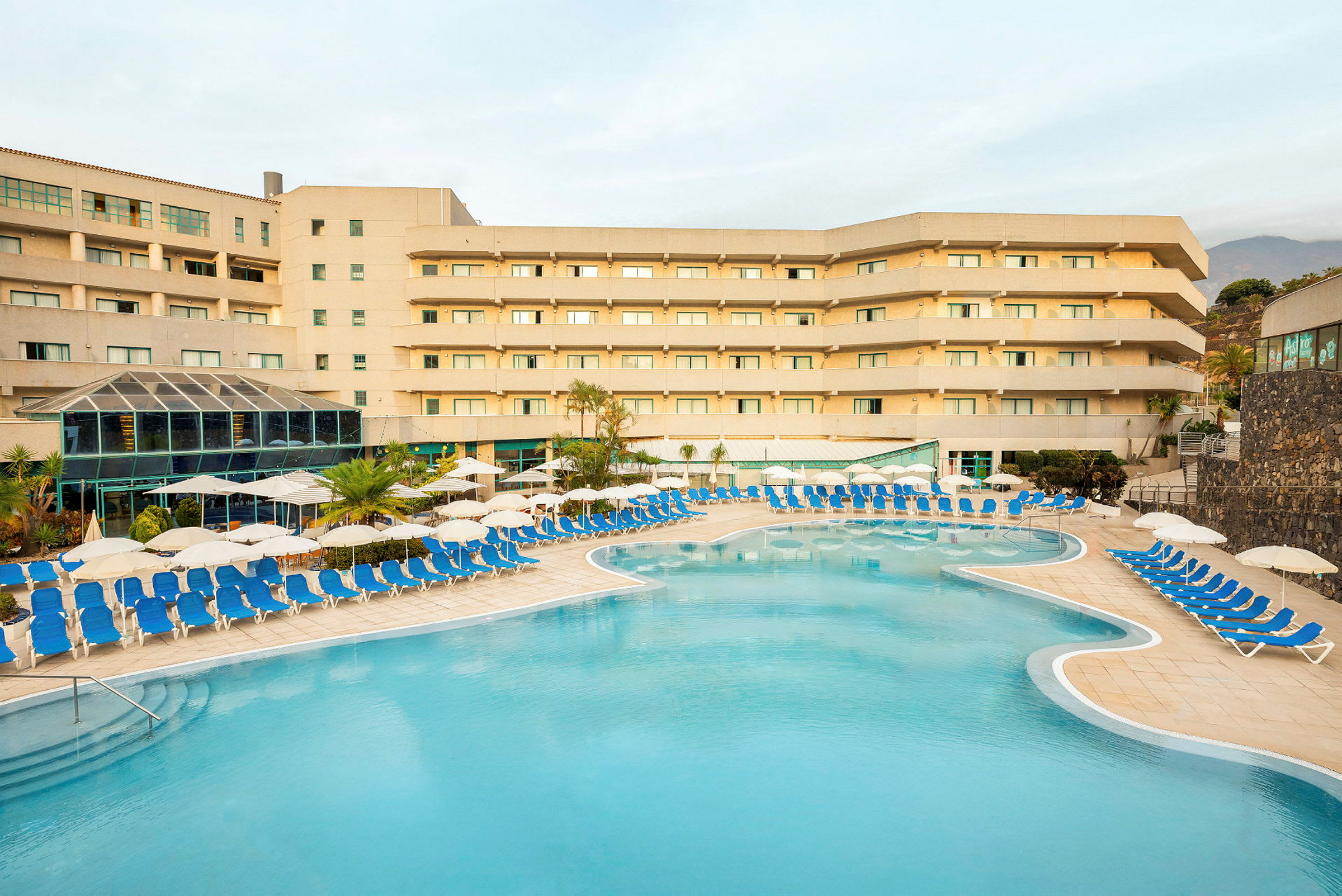 Ett hotellkomplex med en utomhuspool omgiven av solstolar och parasoller. (AI-genererad text)
