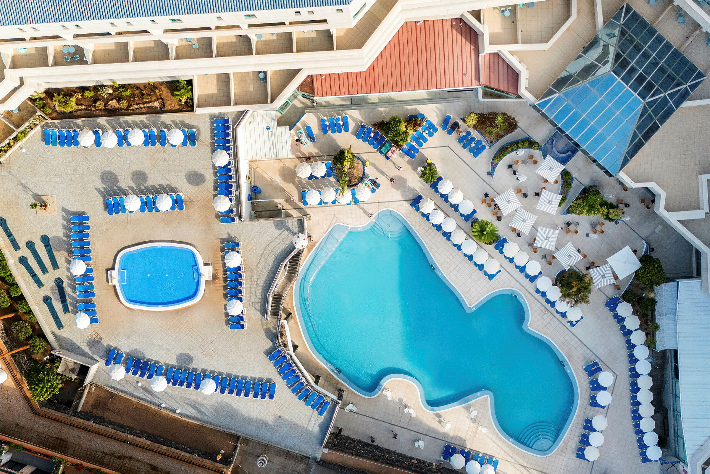 Flygfoto över ett hotellpoolområde med två pooler och solstolar runtom. (AI-genererad text)