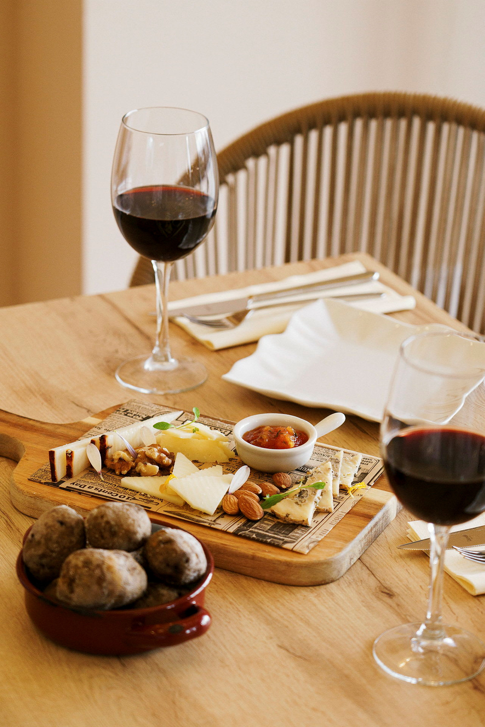 Ett träbord dukat med två vinglas fyllda med rött vin samt en tallrik med olika ostar, nötter, sylt och bröd. (AI-genererad text)