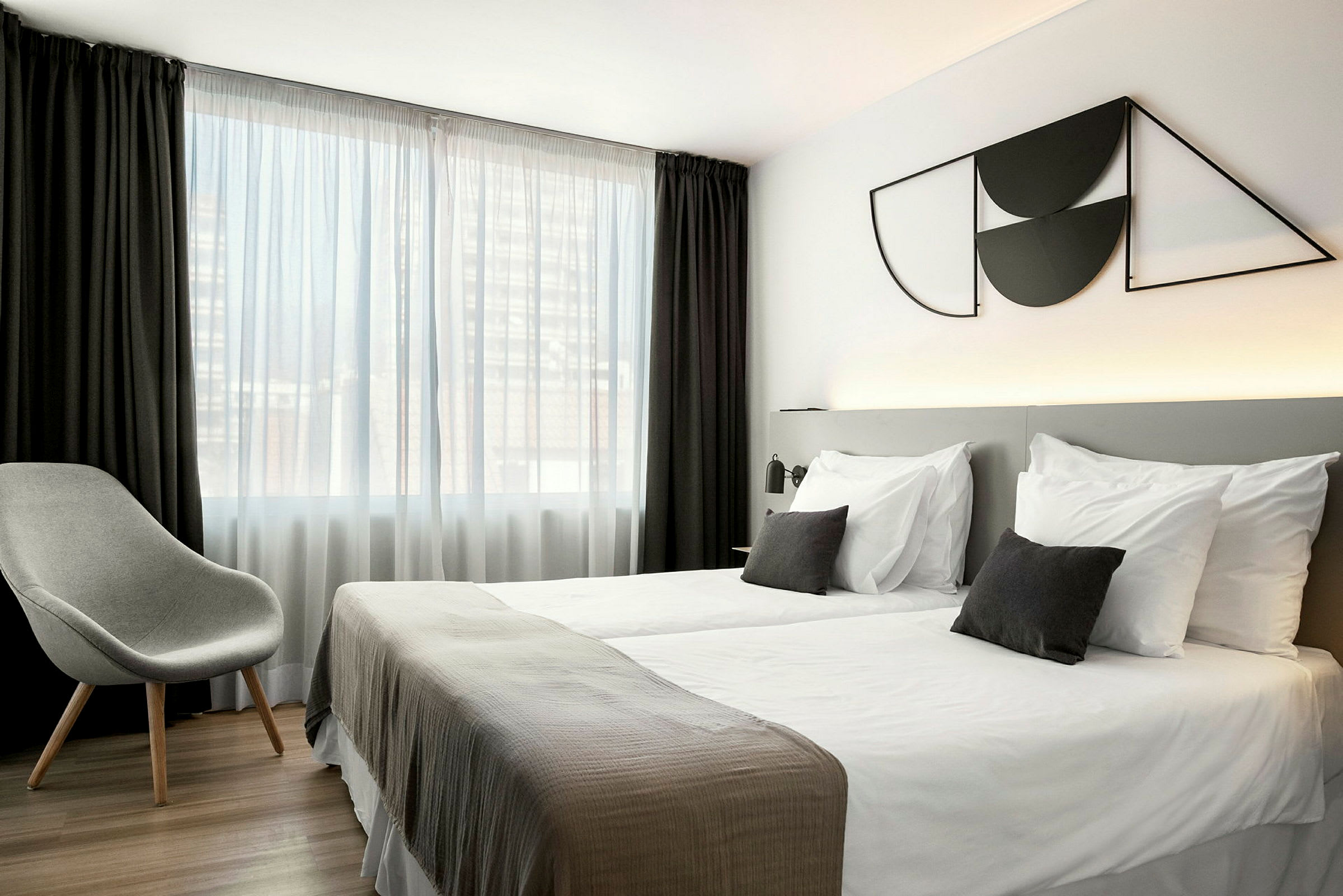 Ett hotellrum med två enkelsängar, en grå stol och ett stort fönster med vita och mörka gardiner. (AI-genererad text)