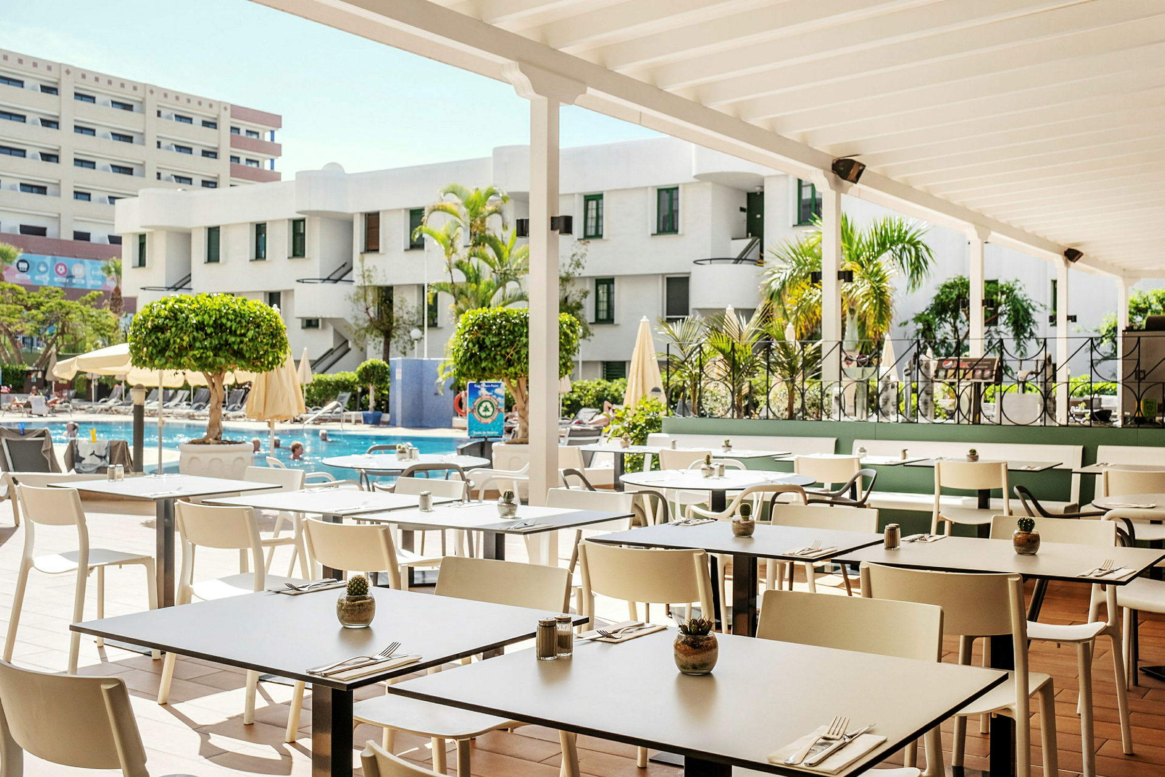 En restaurangterrass med utsikt över poolområdet, omgiven av byggnader och grönska. (AI-genererad text)