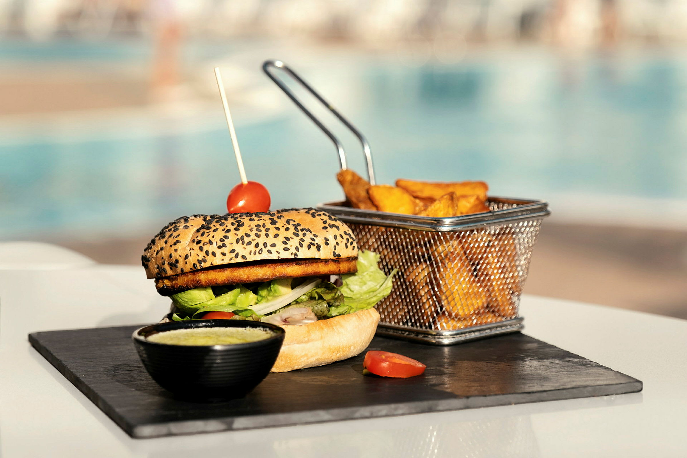En hamburgare med tomatdekoration, vid sidan av pommes frites i en metallkorg, serverad på en stenplatta. (AI-genererad text)