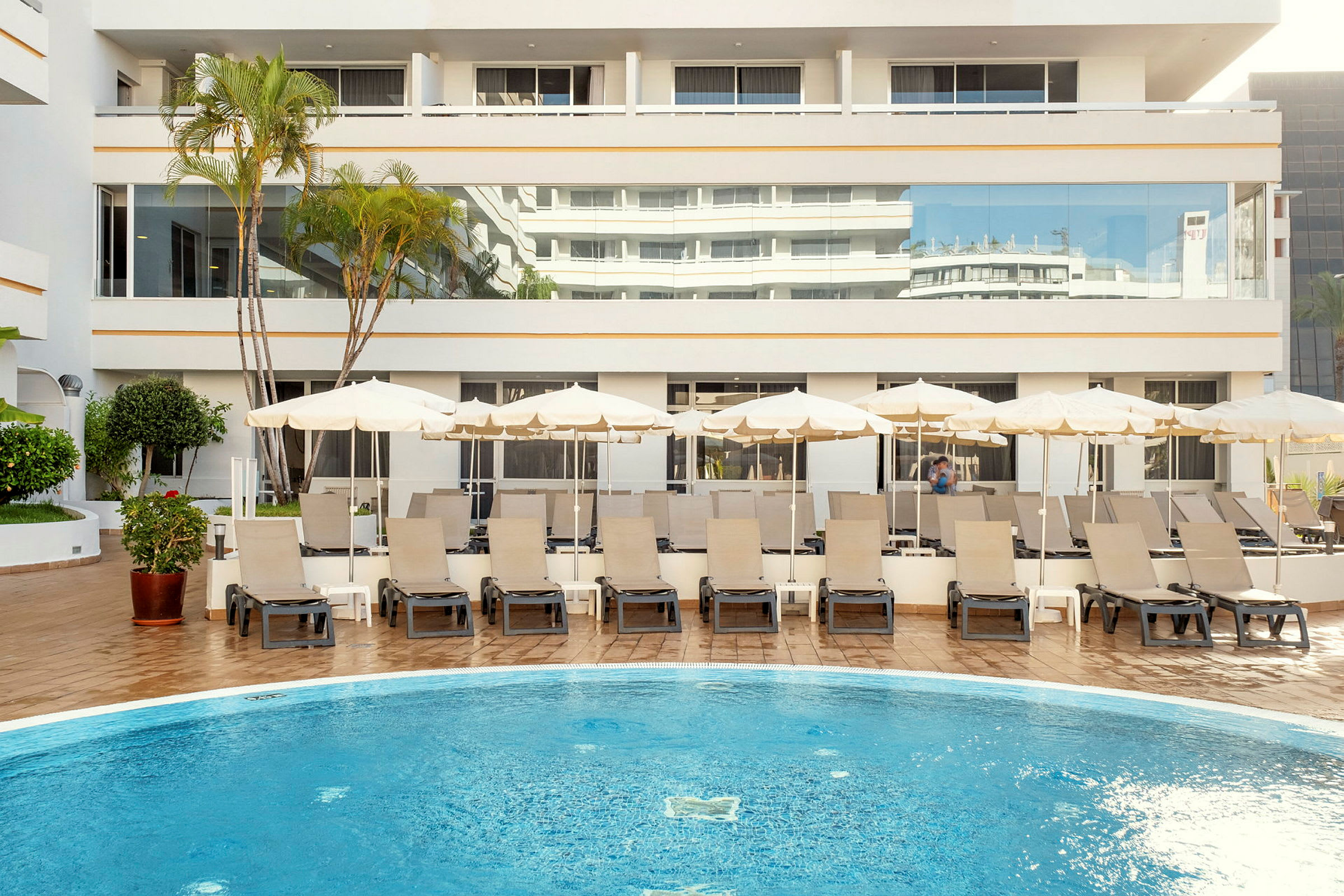 Bild av en hotellpool med solstolar och parasoller framför en ljus byggnad. (AI-genererad text)