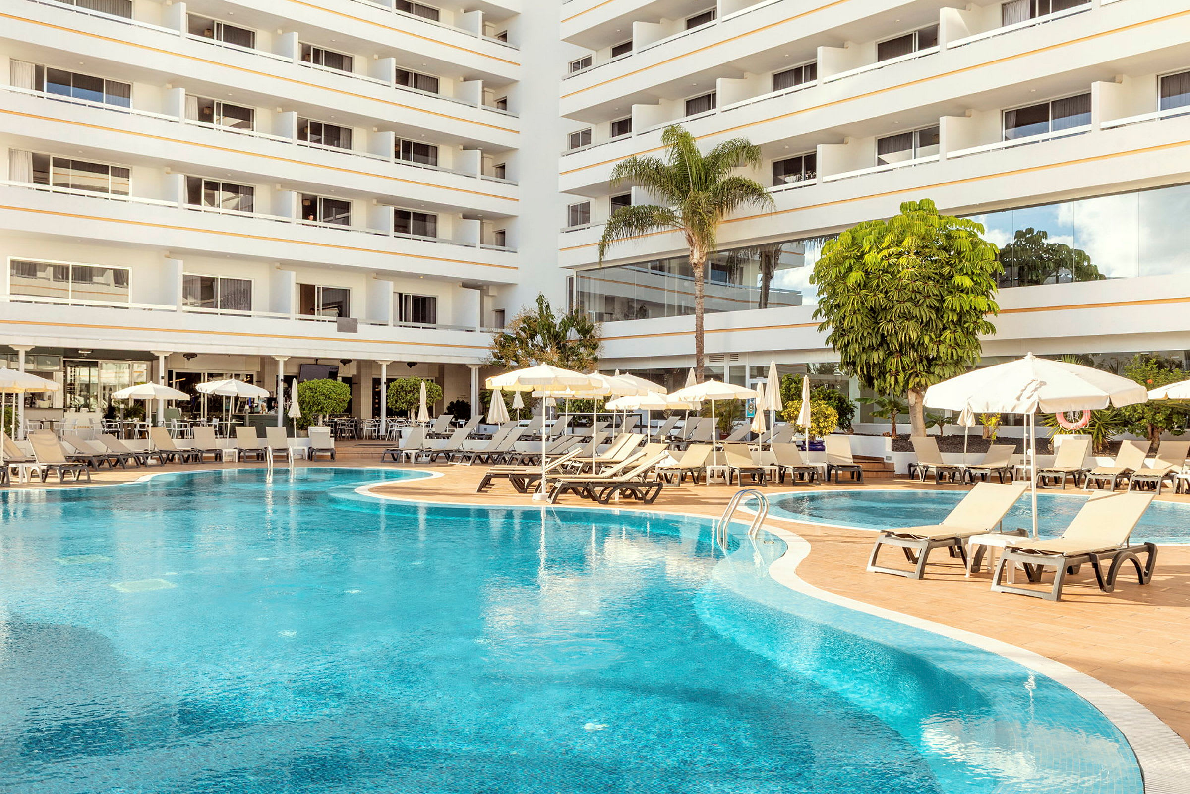 Bild av en hotellpool med solstolar och parasoller vid sidan av och ett fler våningshotell i bakgrunden. (AI-genererad text)