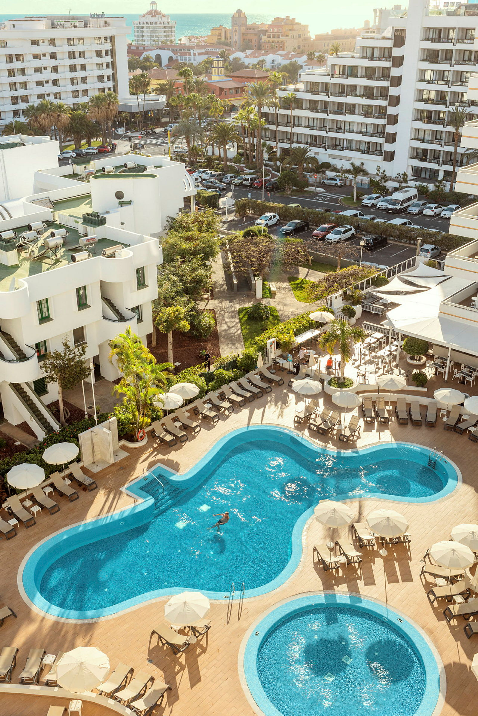 Bild av ett hotellområde med pooler och solstolar, omgiven av gröna växter och byggnader. (AI-genererad text)