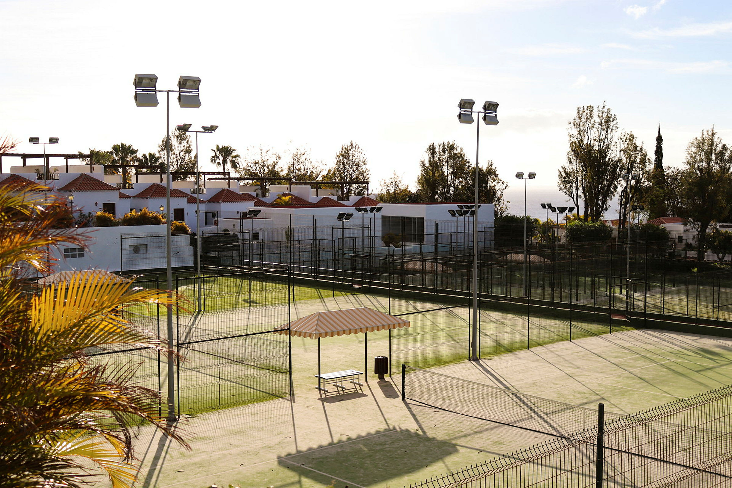 Bild av en paddeltennisbana omgiven av stängsel, med utsikt över en byggnad med vita väggar och röda tak. (AI-genererad text)