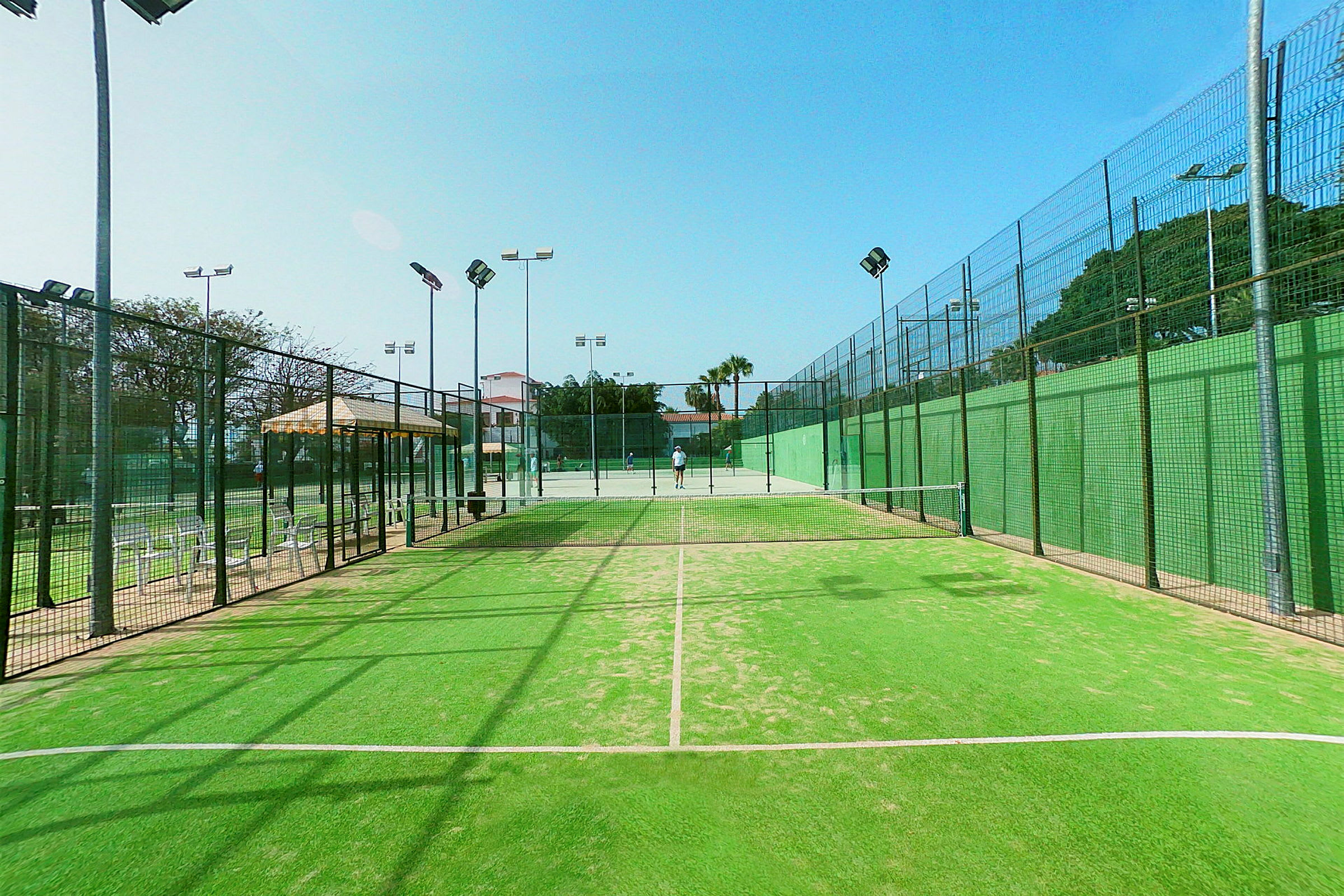 En grön padelbana med nät och belysning, omgiven av staket. (AI-genererad text)