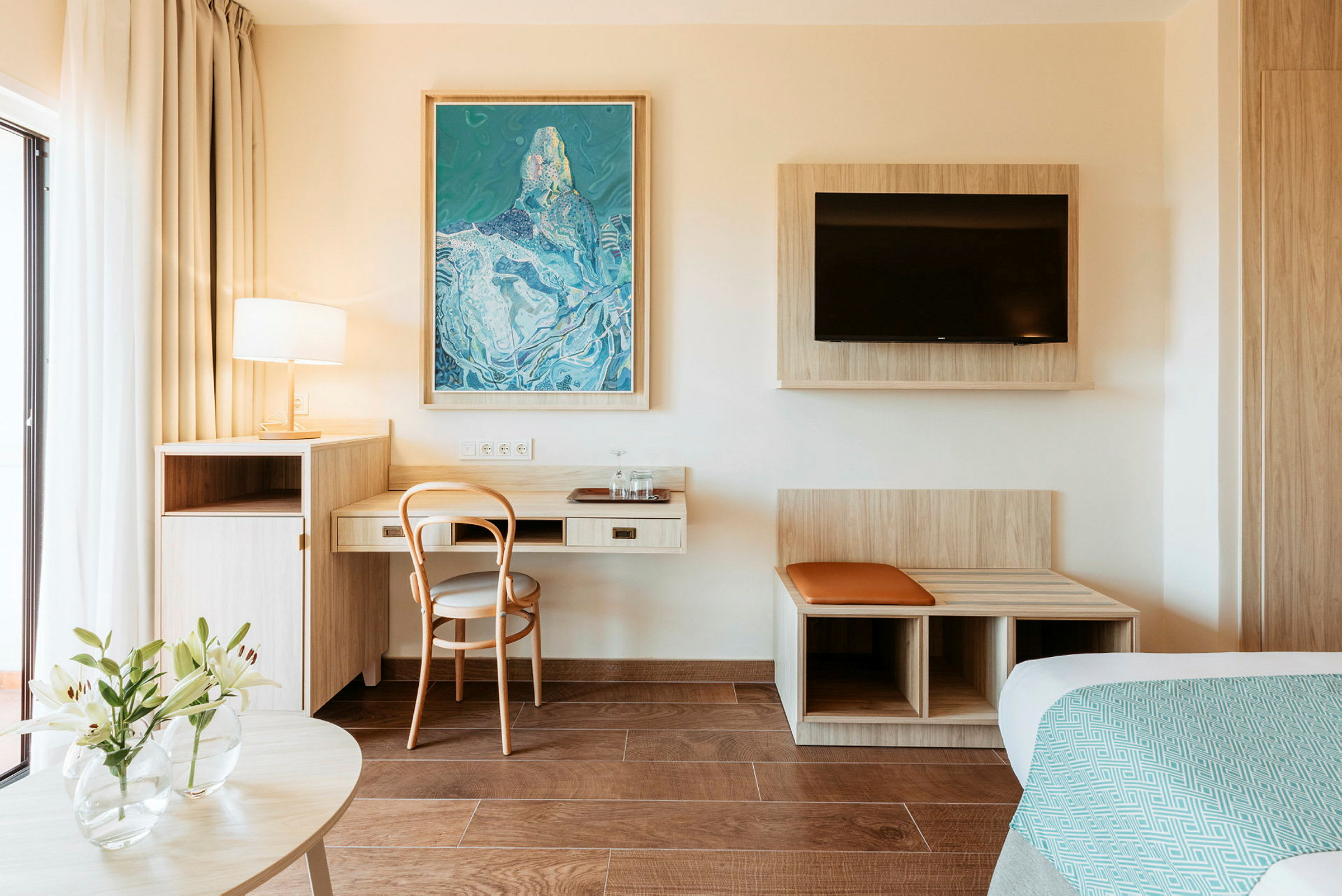 Ett hotellrum med ett skrivbord, en stol, en väggmonterad TV och en dekorativ målning. (AI-genererad text)
