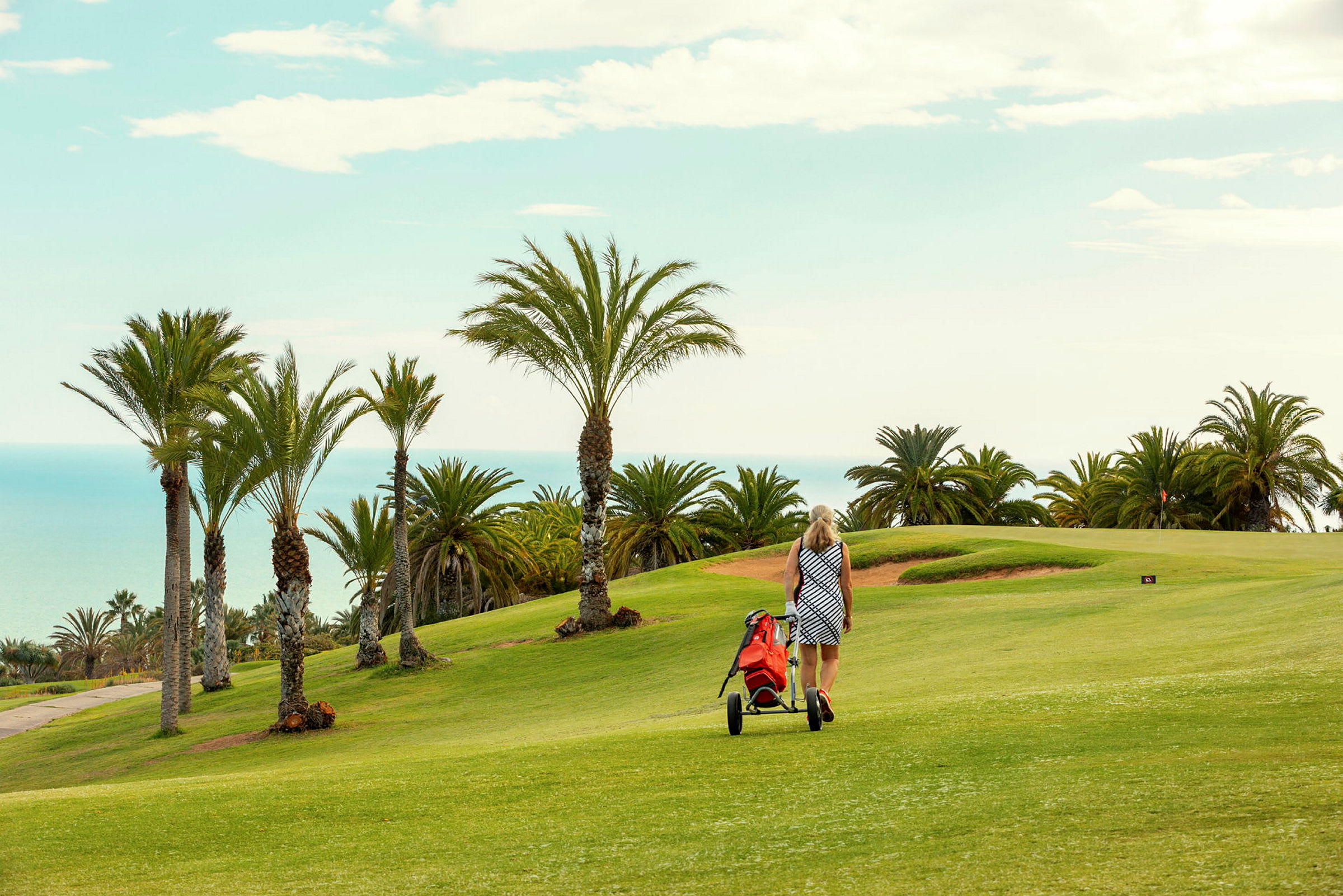 En kvinna går med en golfvagn på en grön golfbana omgivna av palmer med havet i bakgrunden. (AI-genererad text)