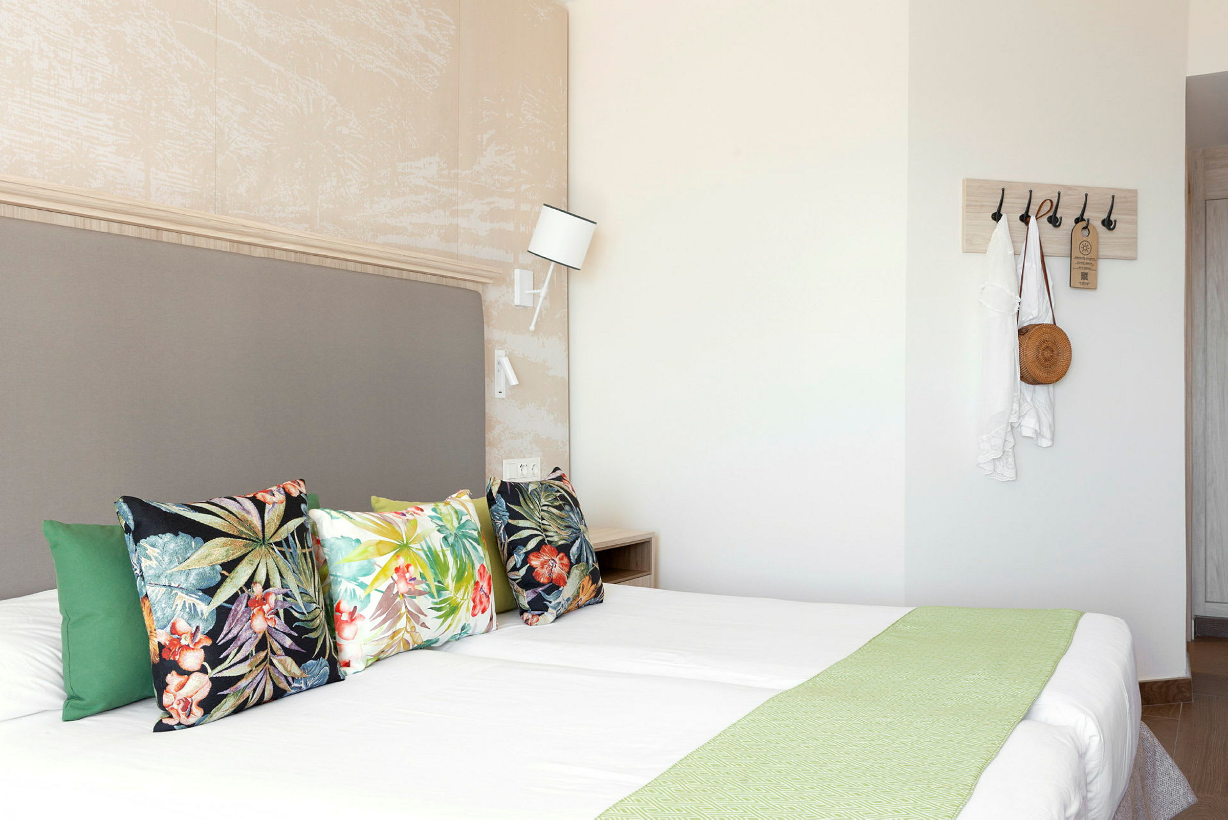 Hotellrum med dubbelsäng, färgglada kuddar och väggkrokar med kläder och väska. (AI-genererad text)