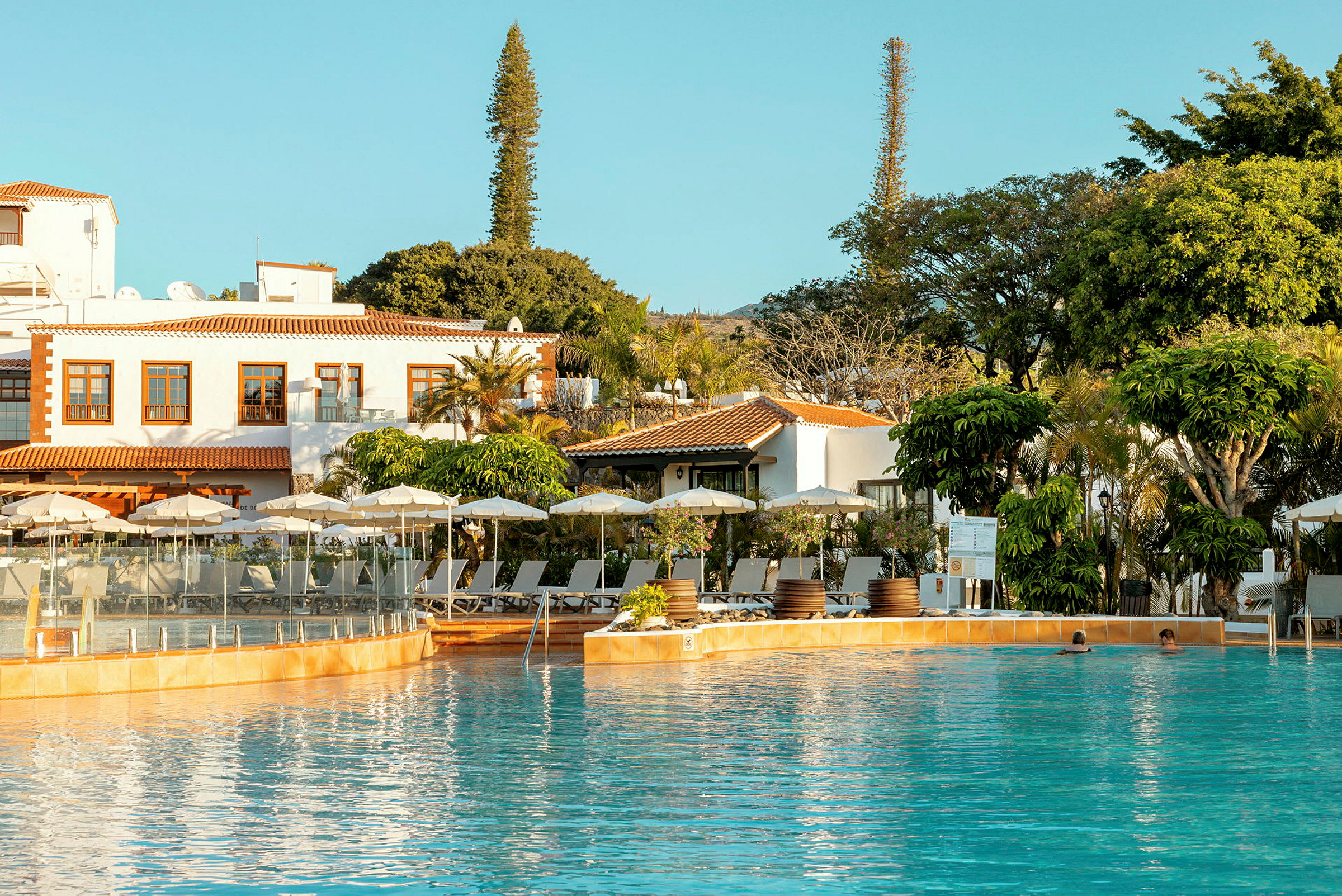 En pool framför ett hotell med parasoller och träd i trädgården. (AI-genererad text)