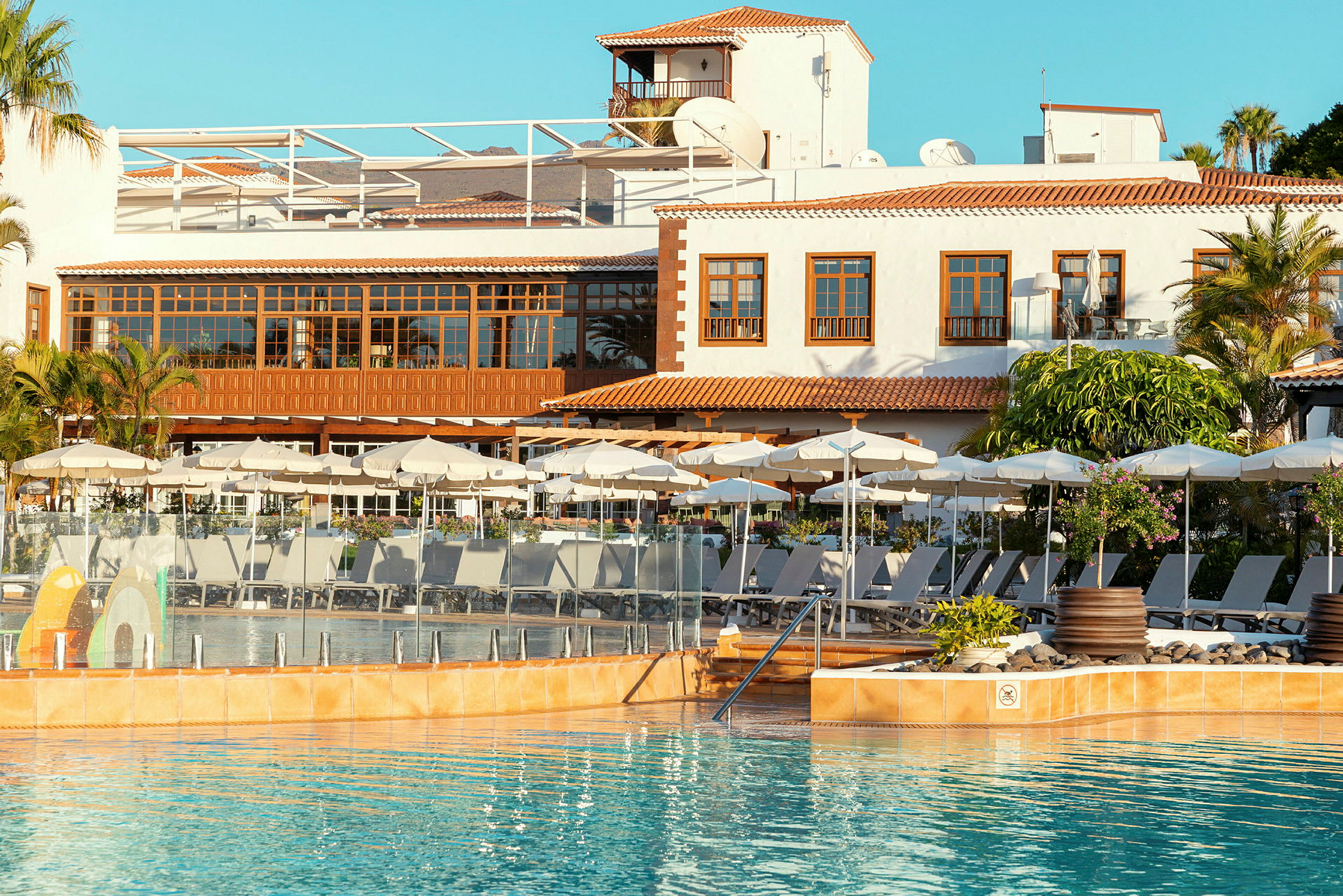 Hotellets poolområde med solstolar och parasoller framför byggnad med terrakottatak. (AI-genererad text)