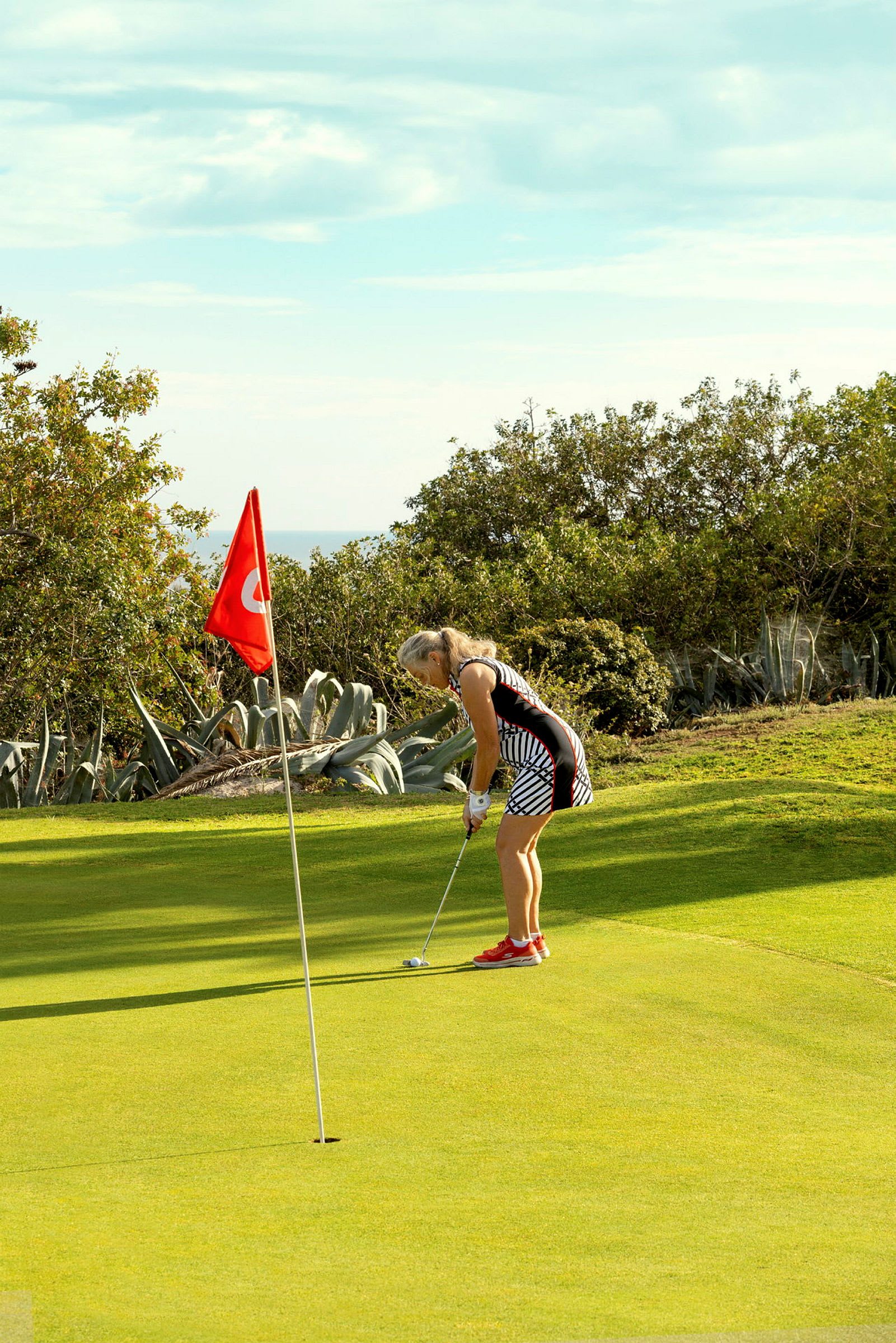 En person spelar golf och siktar på hålet i närheten av en röd flagga, med grön natur i bakgrunden. (AI-genererad text)